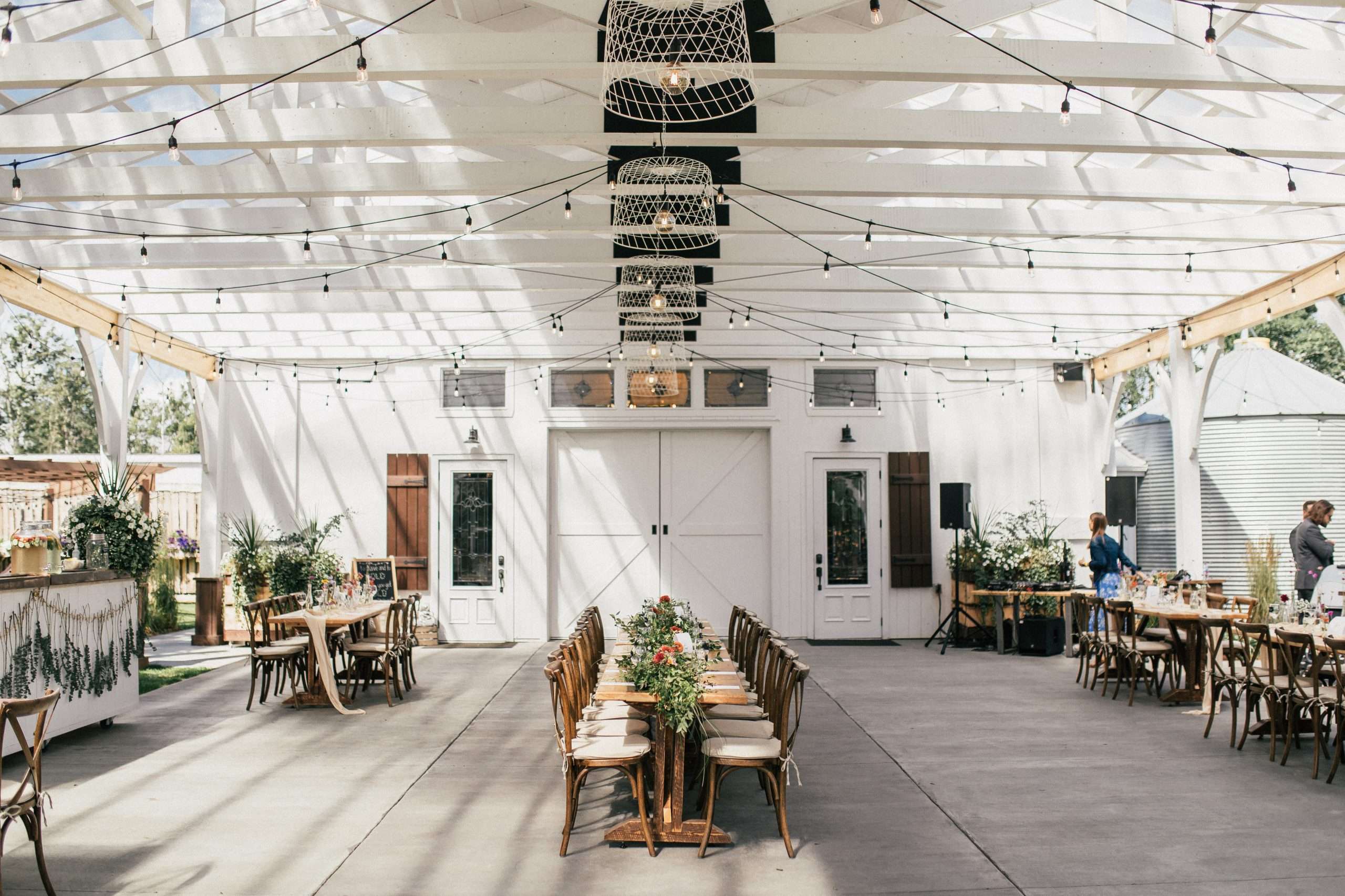 Light-filled barn reception space with long wooden tables, greenery centerpieces, string lights and white rafters.