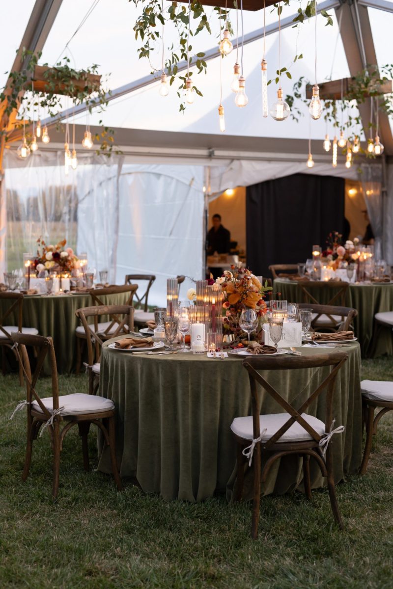 Heritage Ranch fall wedding reception in Bozeman, Montana, featuring green velvet linens, candlelit tables, hanging greenery, Edison bulbs and warm floral centerpieces inside a clear-top tent.