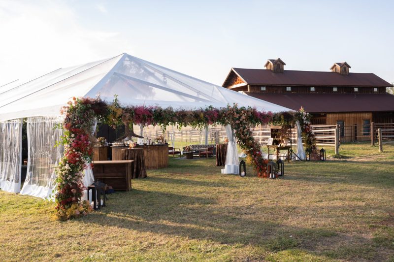 Outdoor fall wedding reception at Heritage Ranch in Bozeman, Montana, with a clear-top tent, colorful floral installation, rustic bar setup and barn backdrop on the ranch property.
