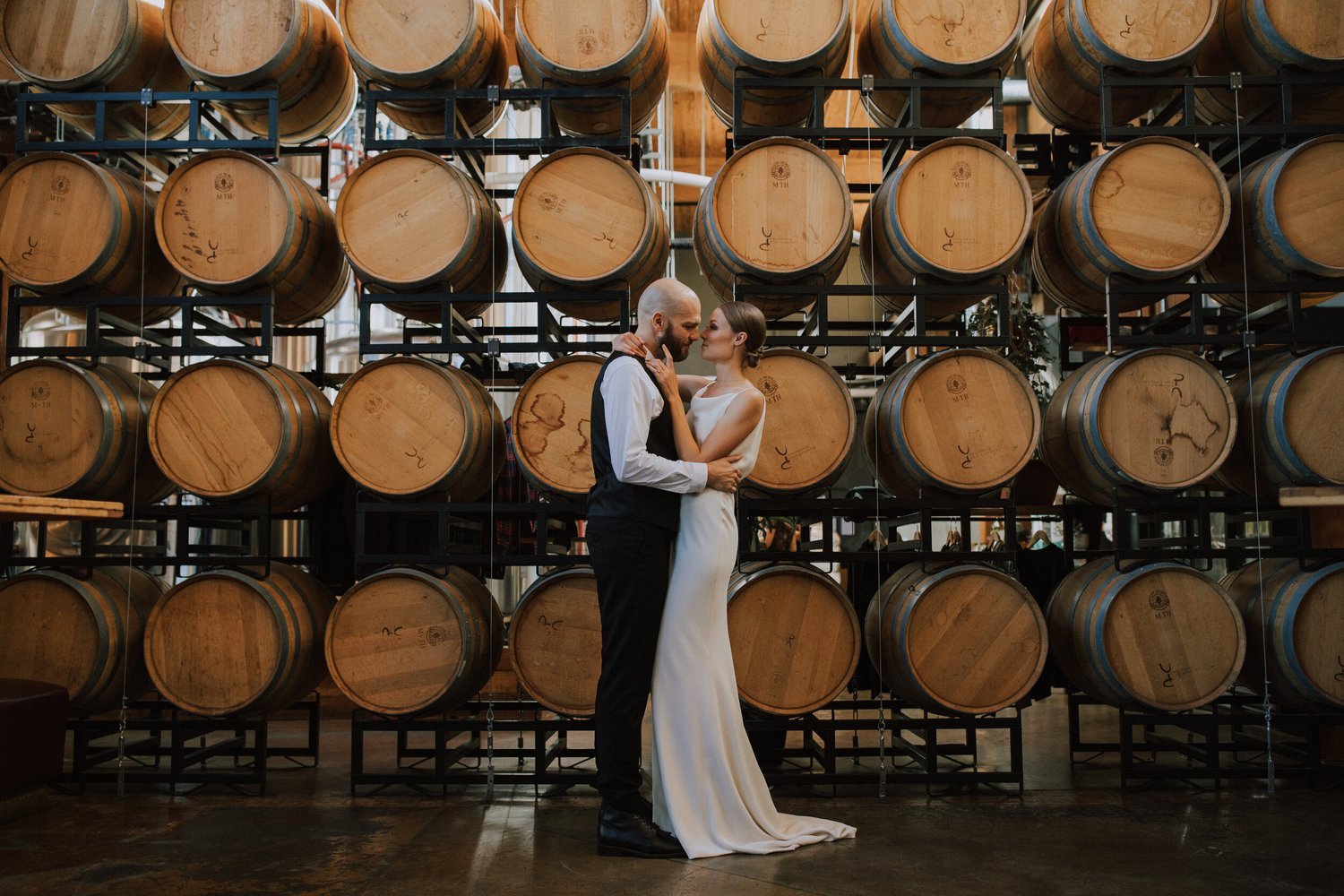 Bride and groom kissing in front of stacked wine barrels at Vancouver Urban Winery.