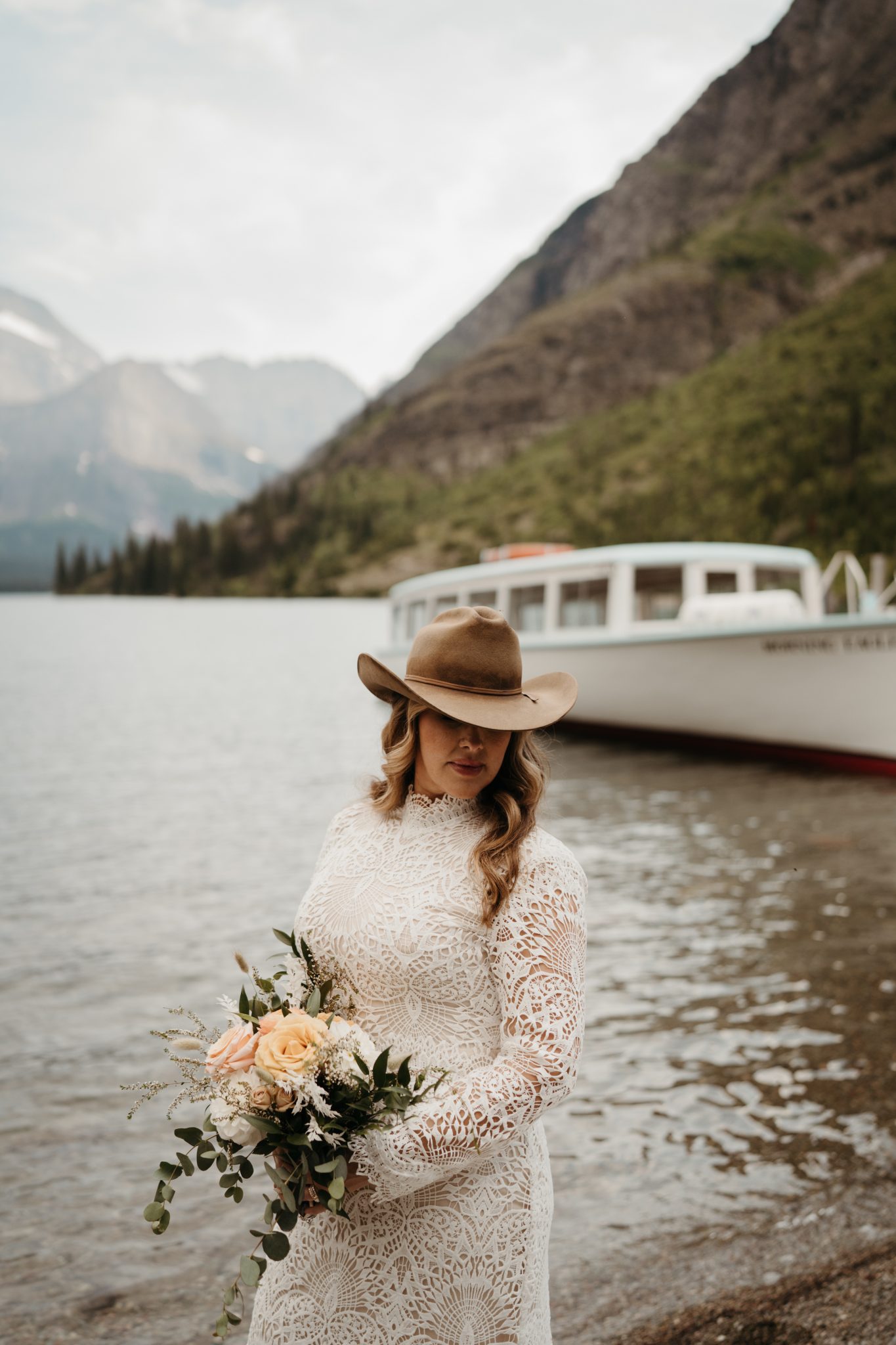 Montana Summer Elopement chic bride with a cowboy hat wearing a long sleeved lace wedding dress while holding her pink and light yellow bouquet with the lake in the background