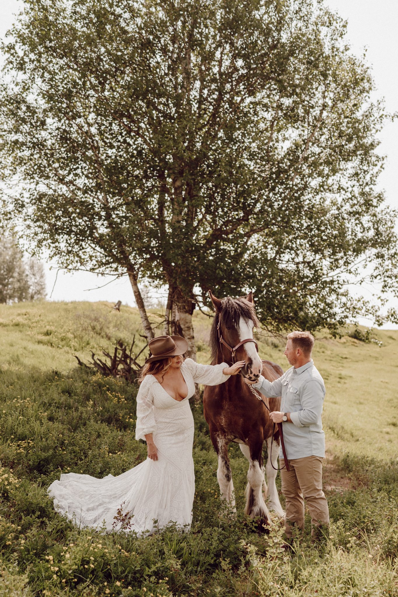Montana Summer Elopement bride and groom petting a horse in a field of grass as she wears a floor length casual white dress and brown cowboy hat and the groom is wearing a blue casual button up and beige pants
