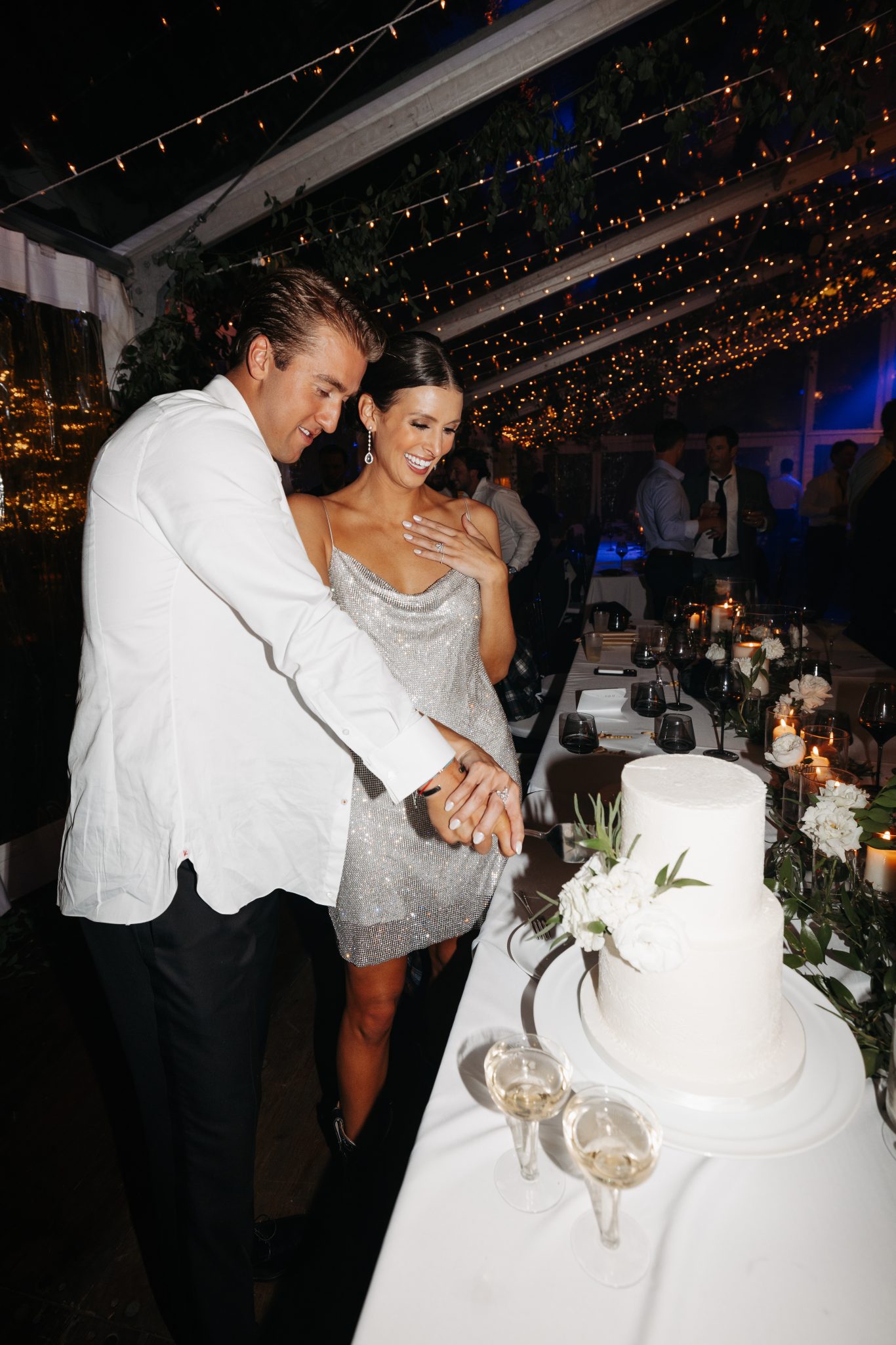 Snowmass Fall Wedding bride and groom cutting into their two-tiered white wedding cake under the twinkling lights in their tent wedding