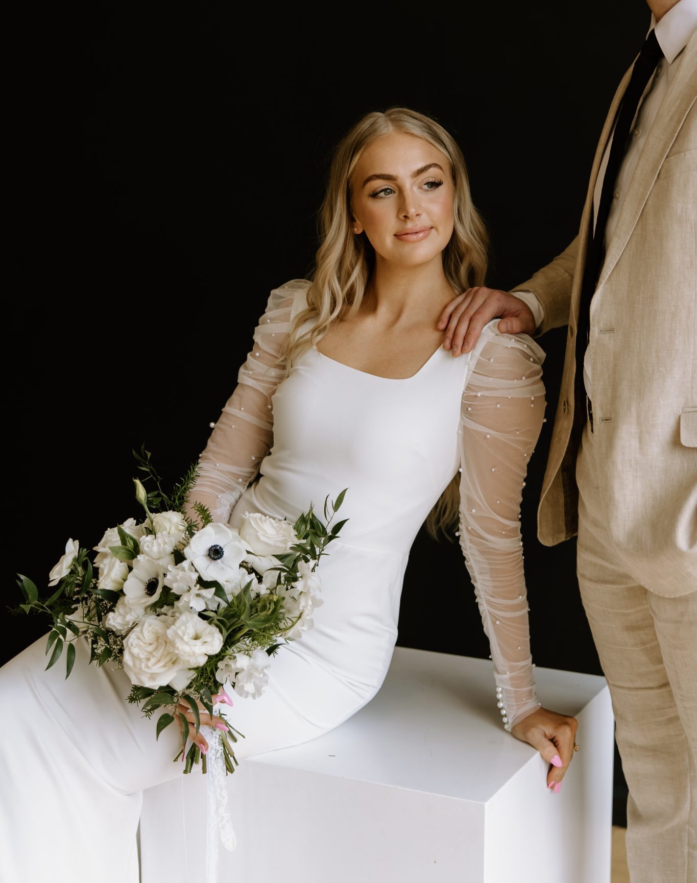 Winter Idaho Bridals at The Paris Studios bride sitting on a white box showing off her fitted wedding dress and illusion mesh sleeves with pearls on them and the groom is resting his arm on her shoulder