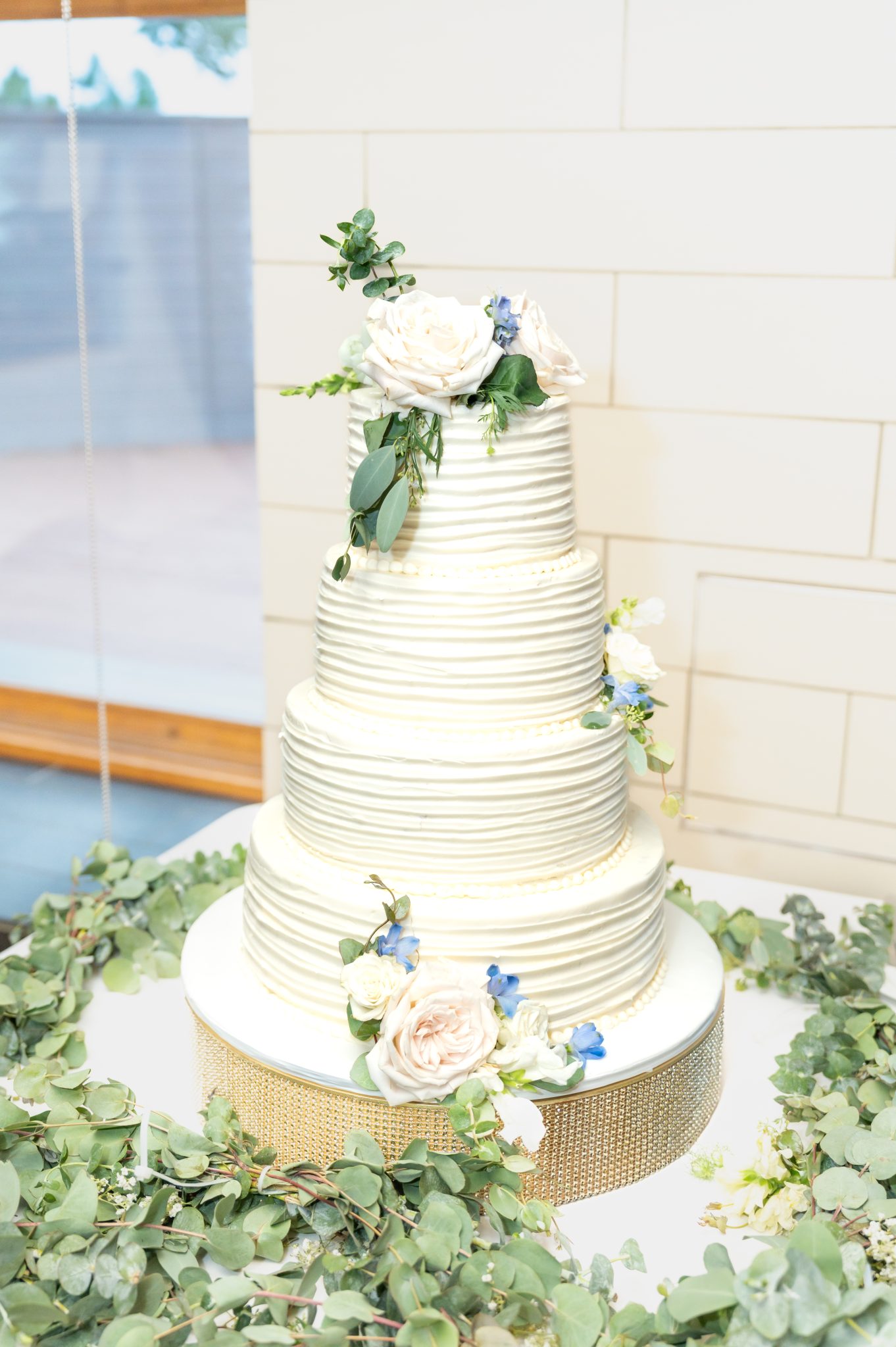 Elegant three-tier wedding cake adorned with white and blue flowers, surrounded by greenery for a Golden summer wedding.