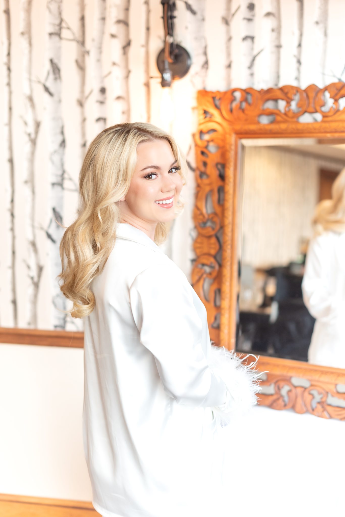 Bride getting ready, smiling in her white bridal robe with feathers, in front of a birch-patterned wall and ornate mirror for her Golden summer wedding.