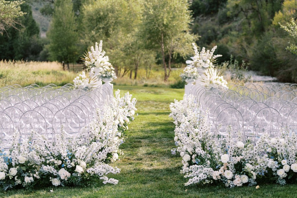 A beautiful outdoor ceremony aisle lined with white flowers and clear chairs at a Peoa Spring Wedding.