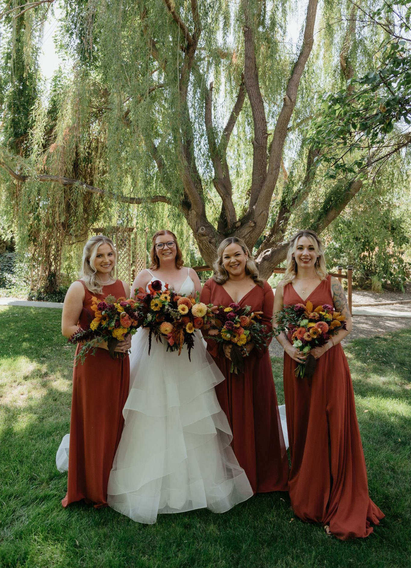 Bride and bridesmaids in matching rust-colored dresses holding colorful bouquets at The Vintage Rose Wedding Venue summer wedding.