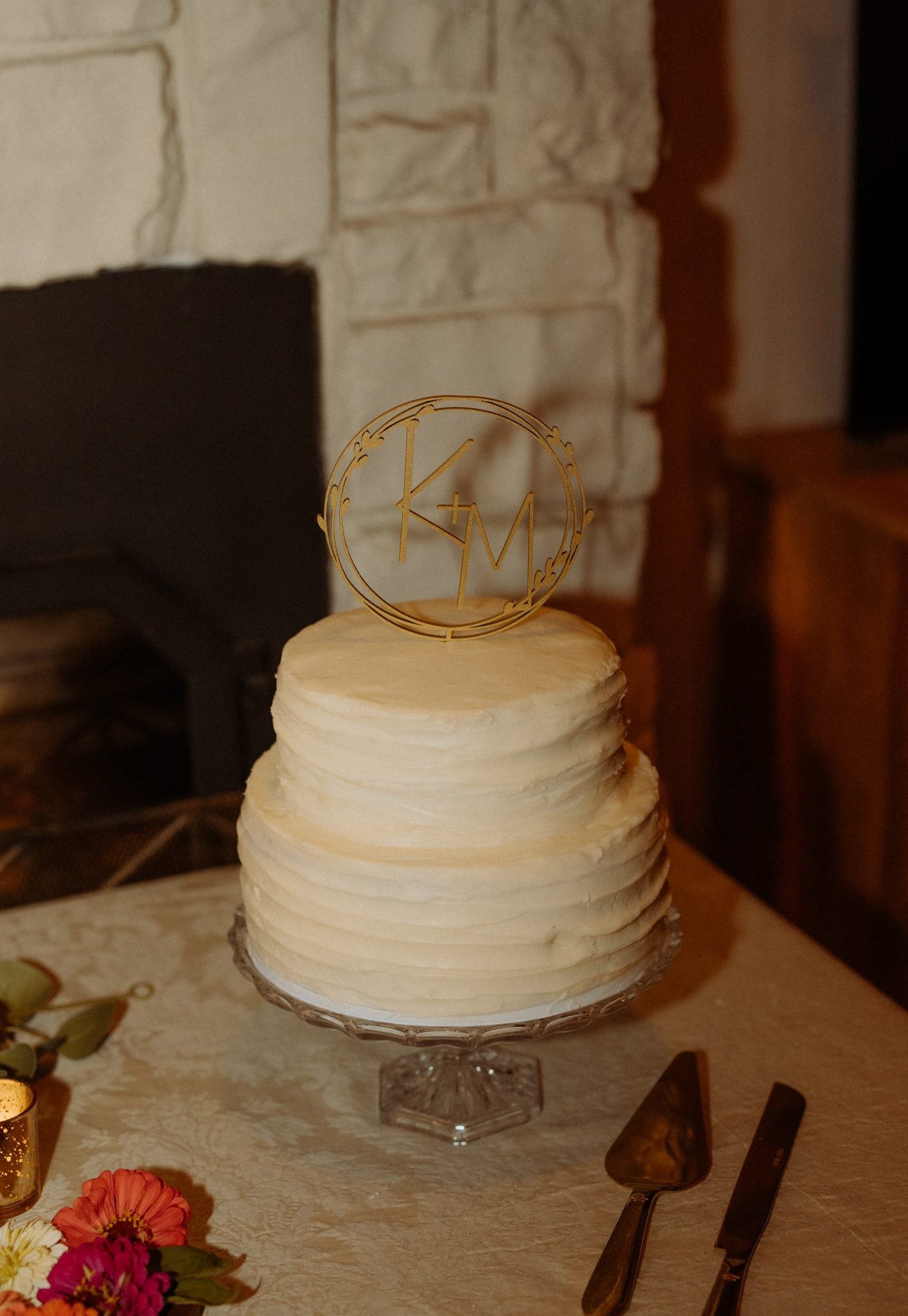 A simple white wedding cake with a custom monogram topper sits on a table, surrounded by flowers, adding a sweet touch to this New Mexico wedding reception.