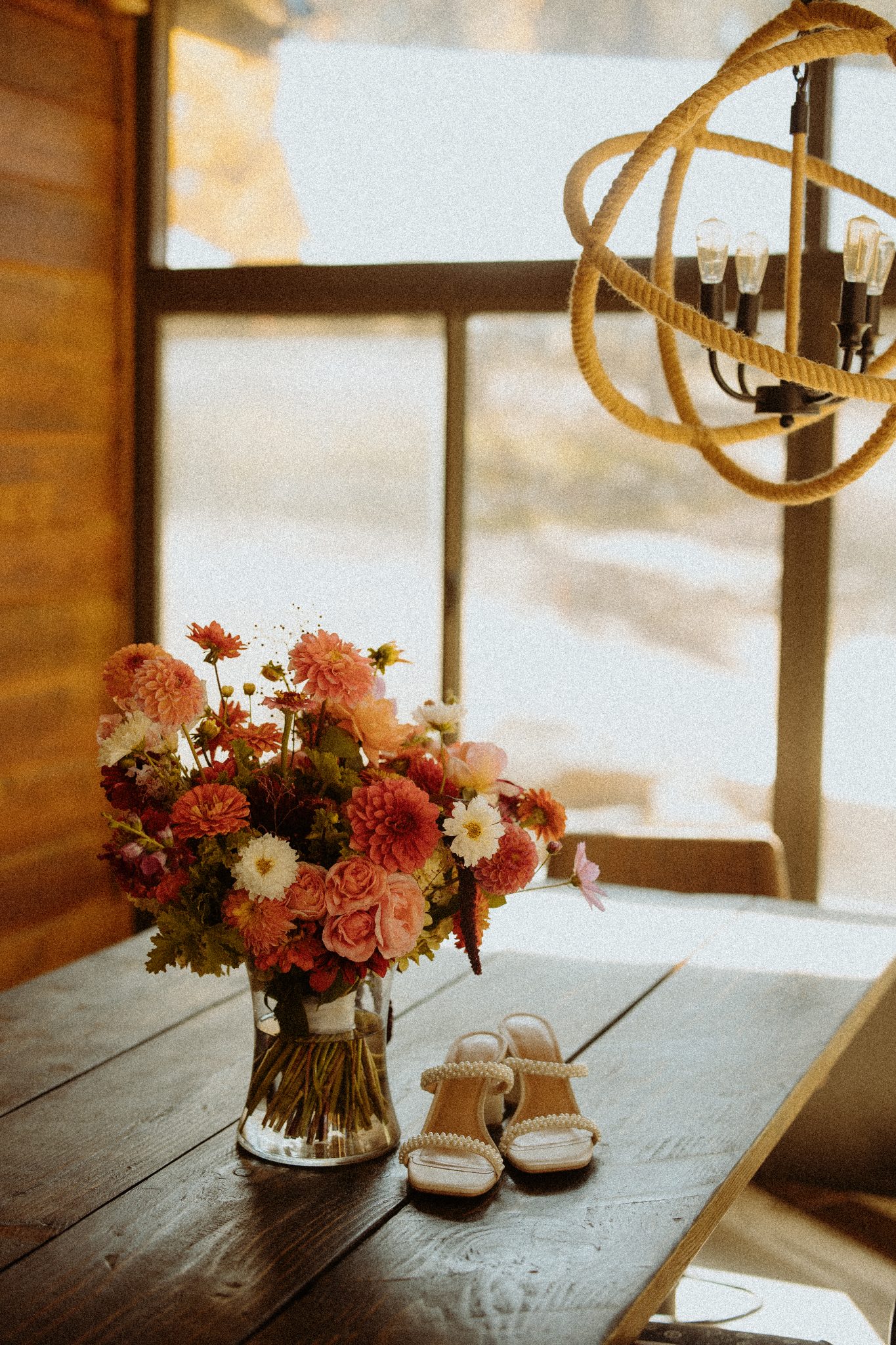 A vibrant bouquet of pink and red flowers sits next to a pair of pearl-embellished shoes on a rustic wooden table, perfect for a New Mexico wedding.