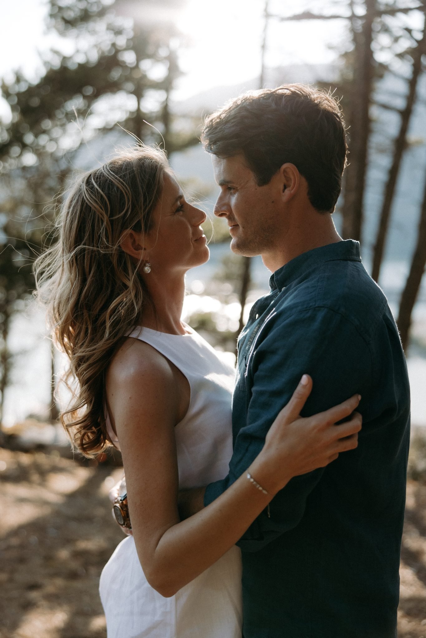 An engaged couple embraces in a forest setting in British Columbia, with sunlight streaming through the trees.