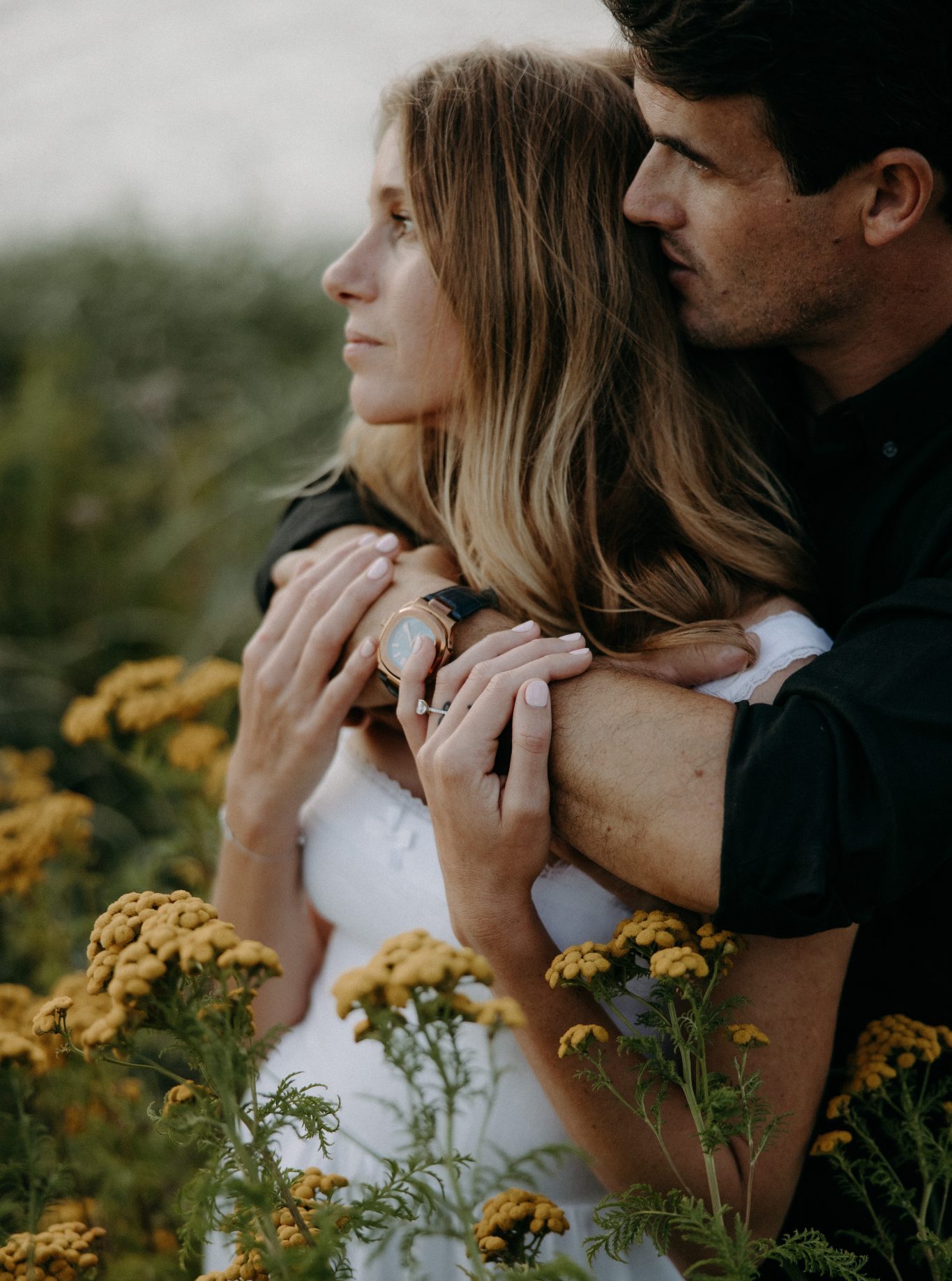 A man tenderly hugs his fiancée from behind as they stand amidst wildflowers during their British Columbia engagement session.
