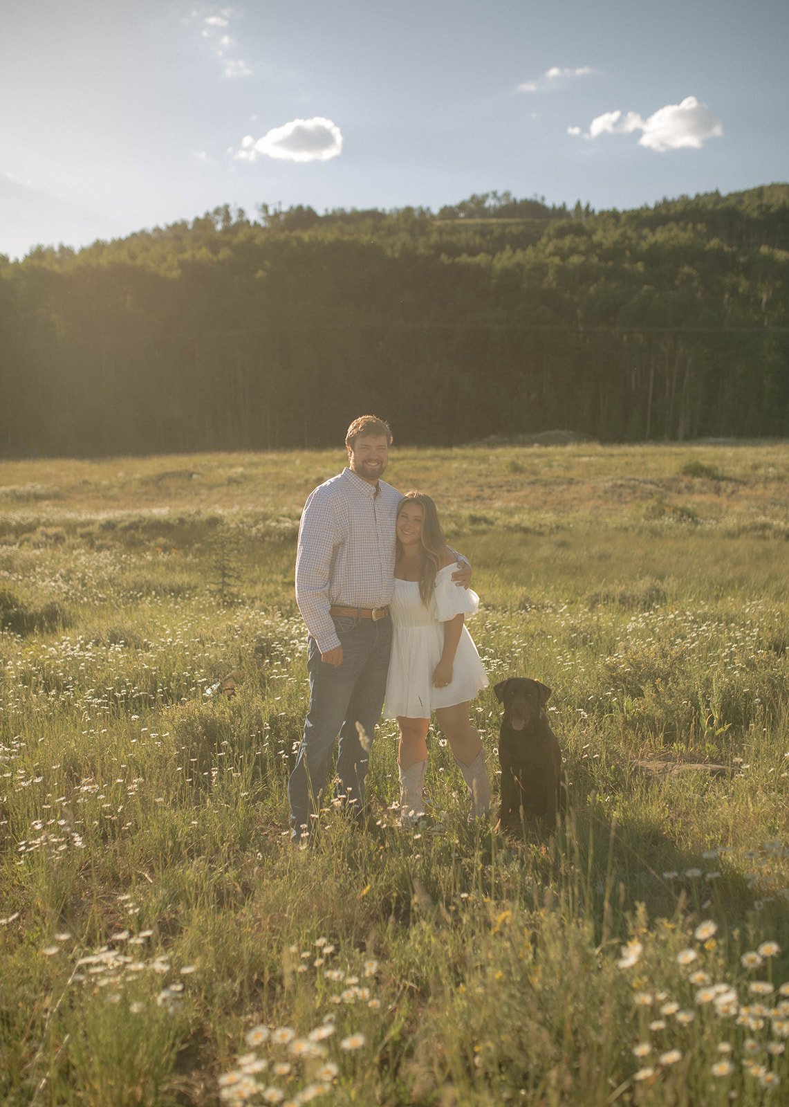 A couple enjoying a romantic moment with their dog during their Durango engagement session in a sunlit meadow.