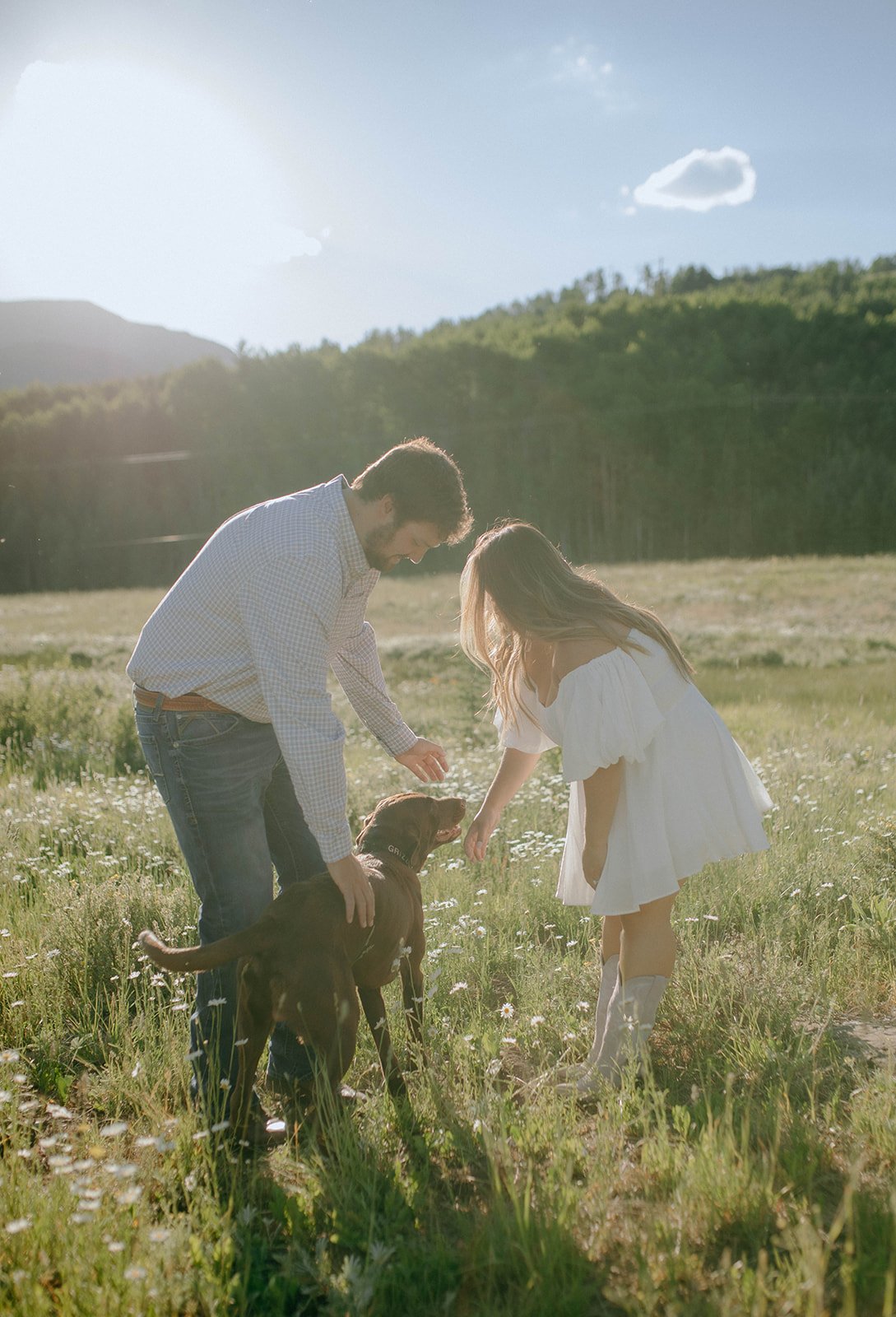 A couple enjoying a romantic moment with their dog during their Durango engagement session in a sunlit meadow.