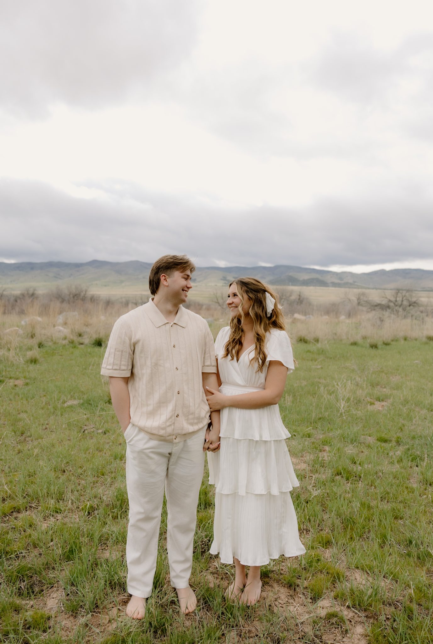 T Beebe Photo Engagement Session at a serene lake, with the couple standing close together in light-colored outfits, enjoying a peaceful moment by the water.