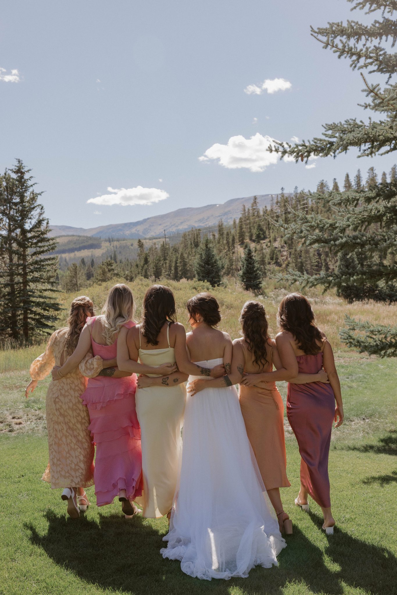 The bride and her bridesmaids in colorful dresses embrace, overlooking the scenic mountains of Breckenridge during a summer wedding.