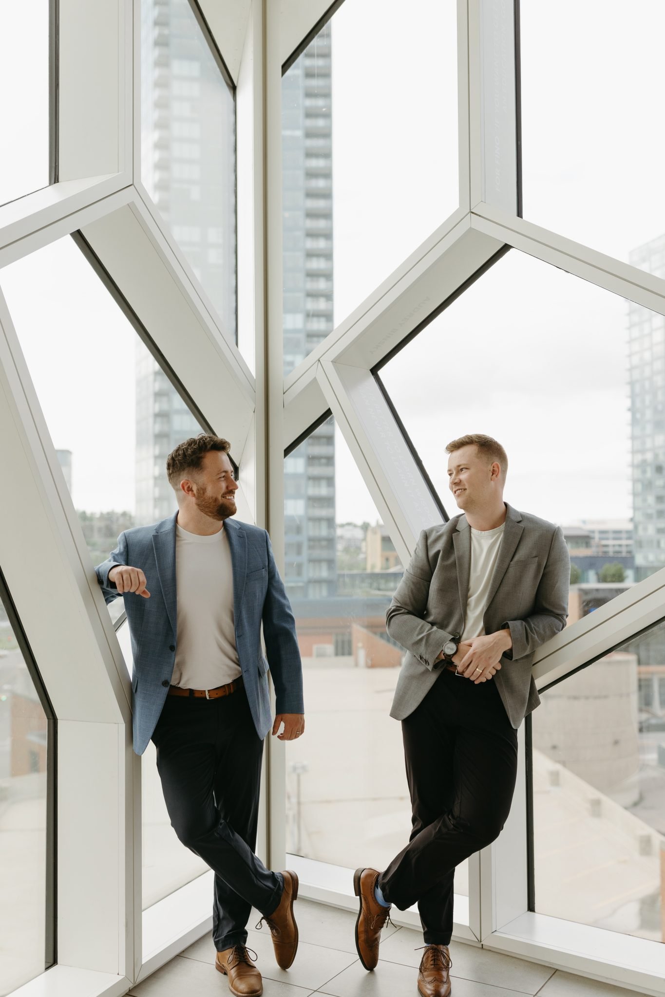 Couple leaning against a geometric window with city views during their cozy Alberta engagement shoot.