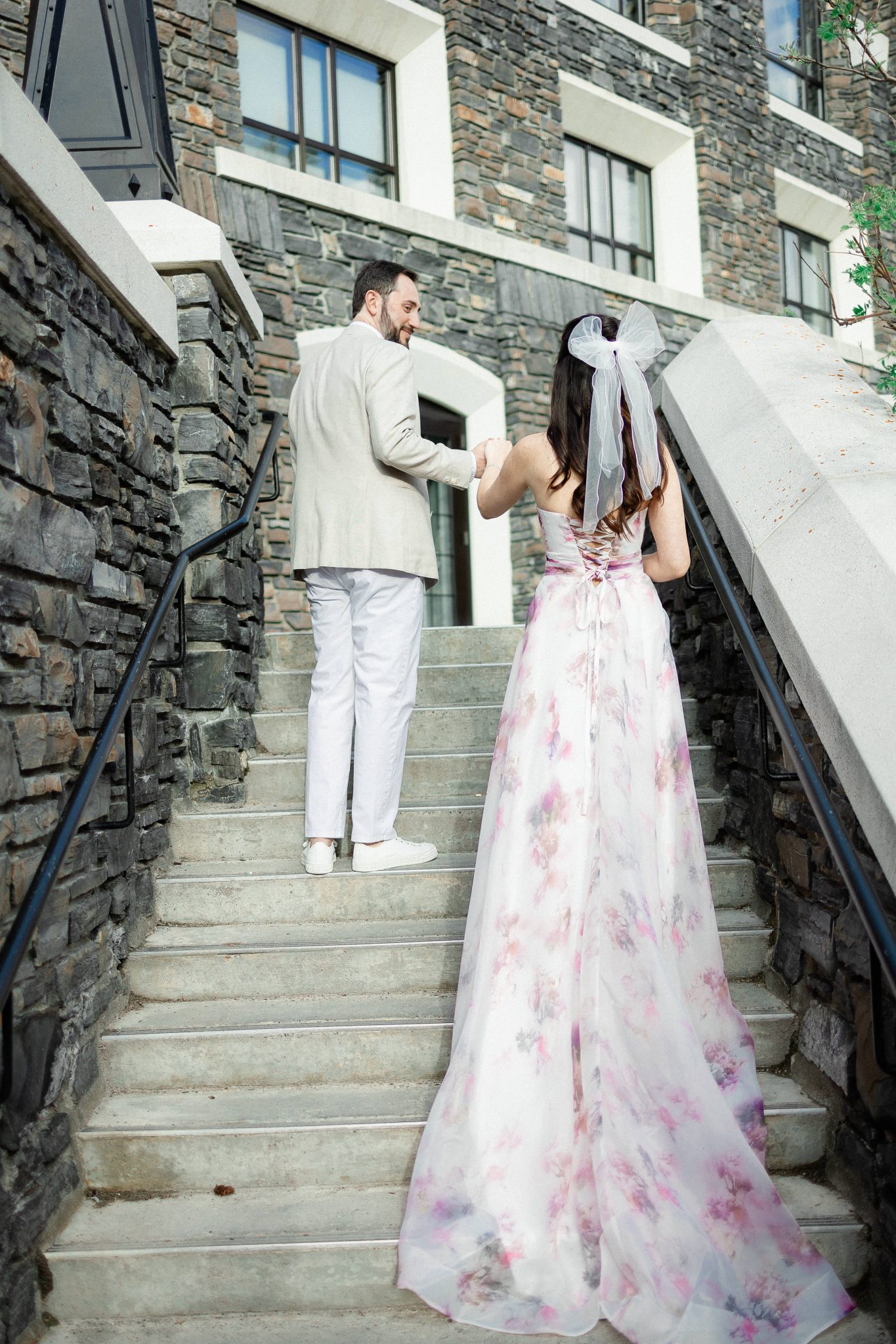 A couple holds hands while walking up a stone staircase, the bride-to-be wearing a flowy floral gown, creating a romantic moment during their soft Alberta engagement session.