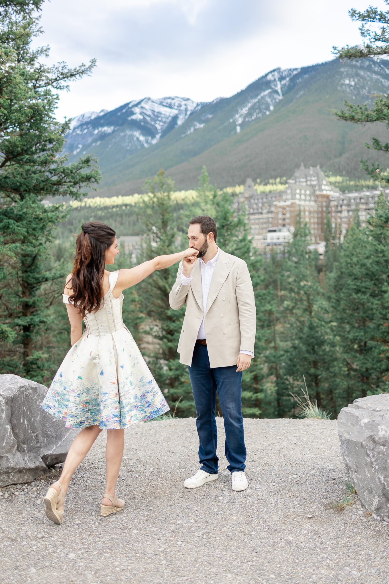 A sweet moment of the couple sharing a kiss, with lush forest greenery and a majestic mountain lodge in the distance, creating an enchanting scene in this Alberta engagement session.
