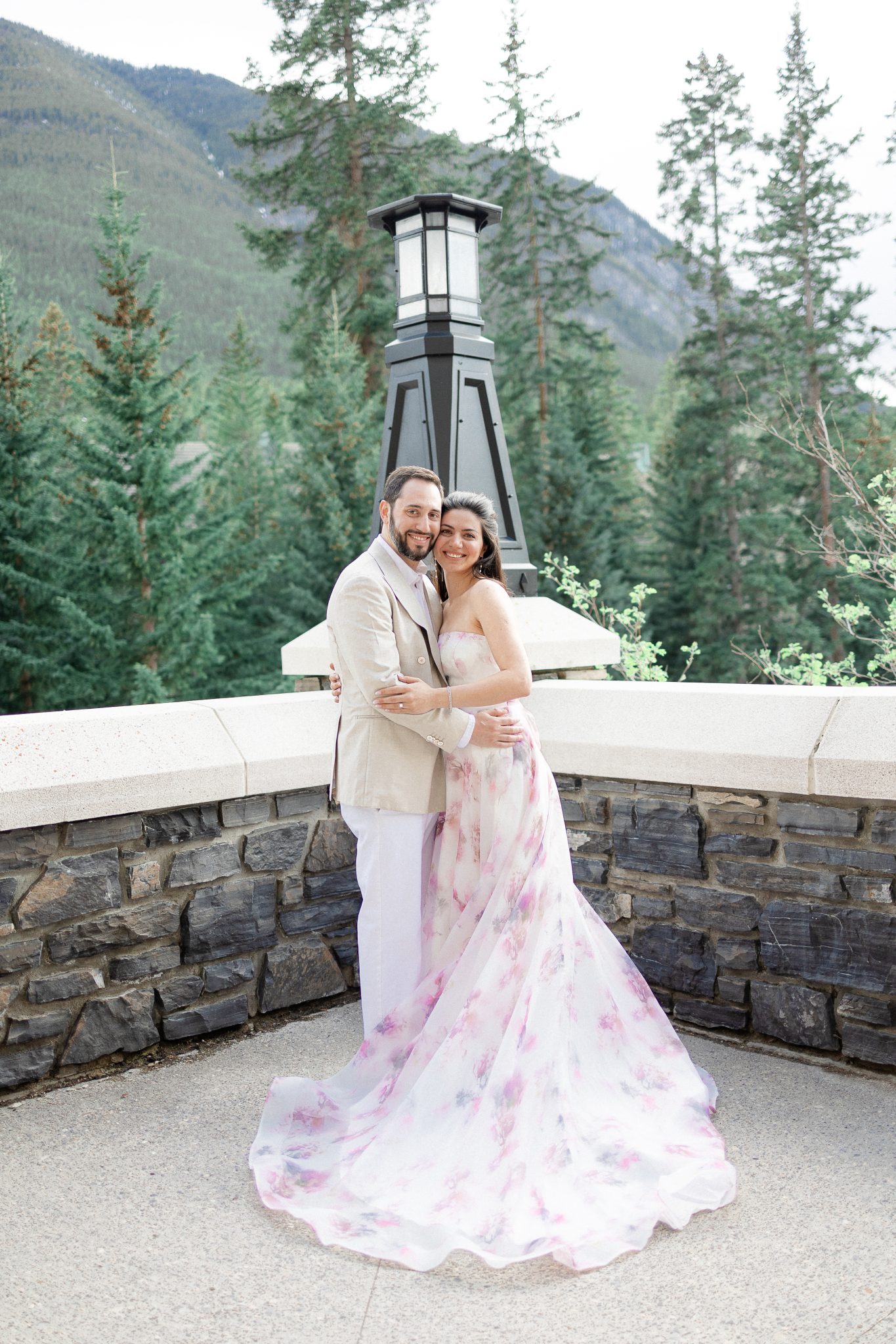 The couple stands together, smiling and embracing against a stunning backdrop of evergreen trees and mountains, perfectly capturing the serene vibe of their Alberta engagement.