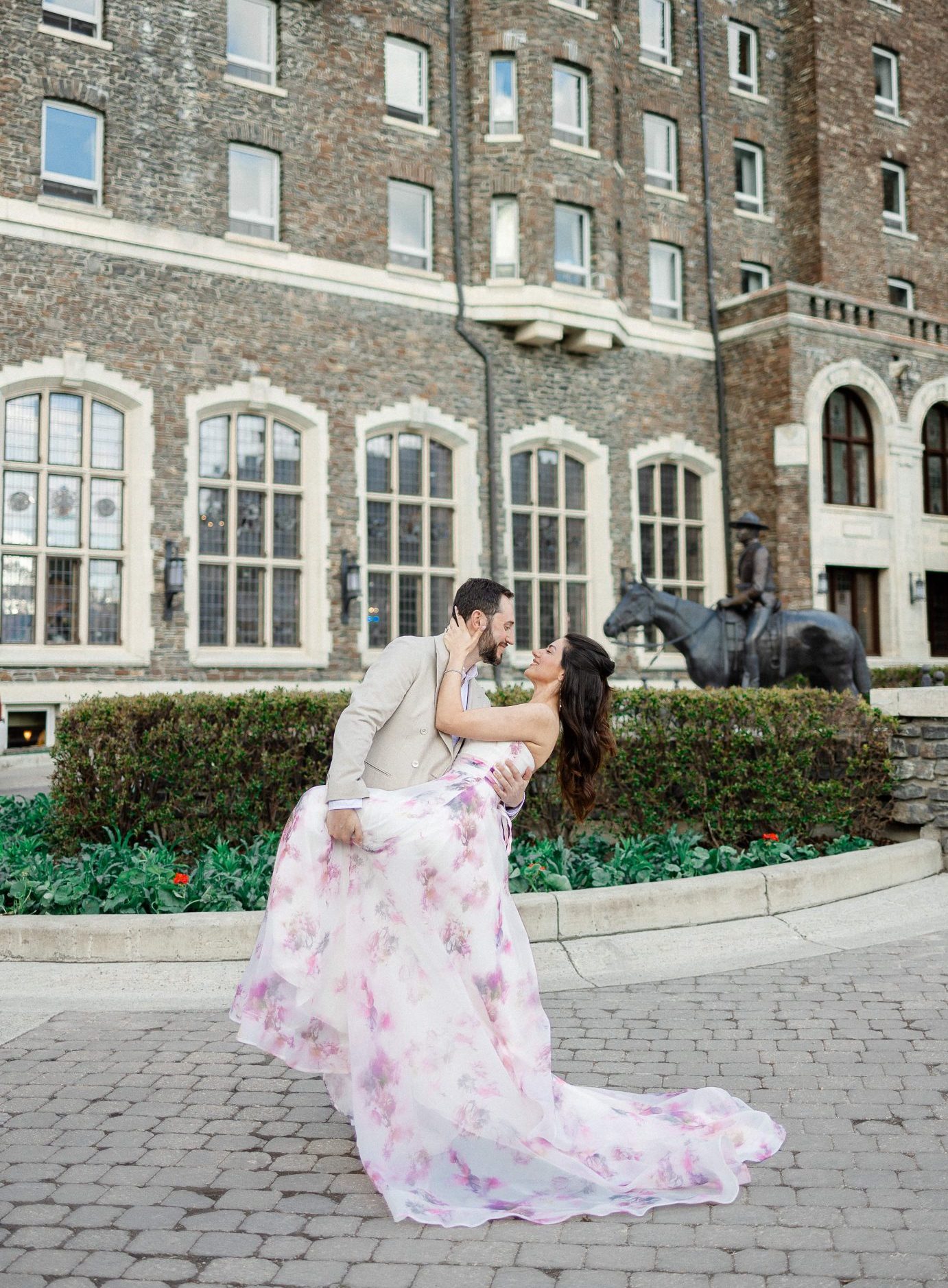A playful dip kiss in front of a historic stone building, with the bride-to-be's pastel floral gown elegantly flowing around her, adding a touch of romance to this Alberta engagement session.