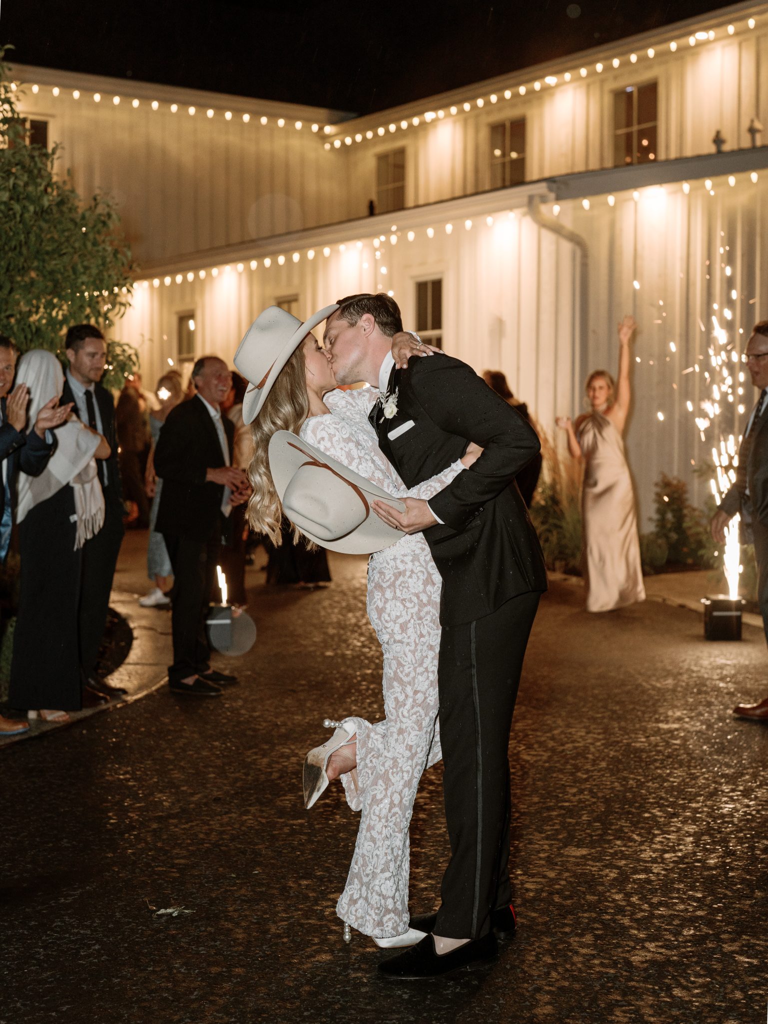 A couple shares a romantic kiss under the night sky, both wearing stylish cowboy hats, surrounded by guests at their summer wedding in Utah.