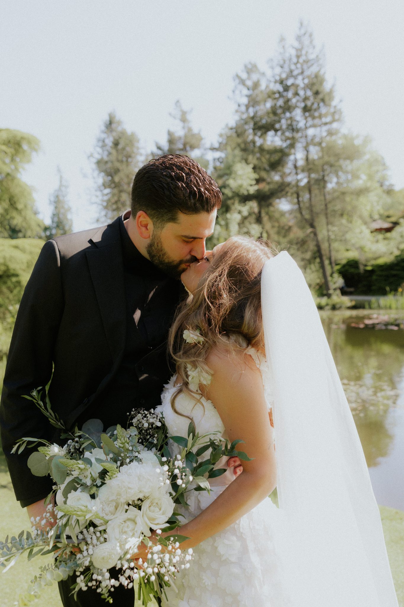 Bride and groom sharing a kiss by the pond, bride holding a white floral bouquet at a Black and White Vancouver Wedding.