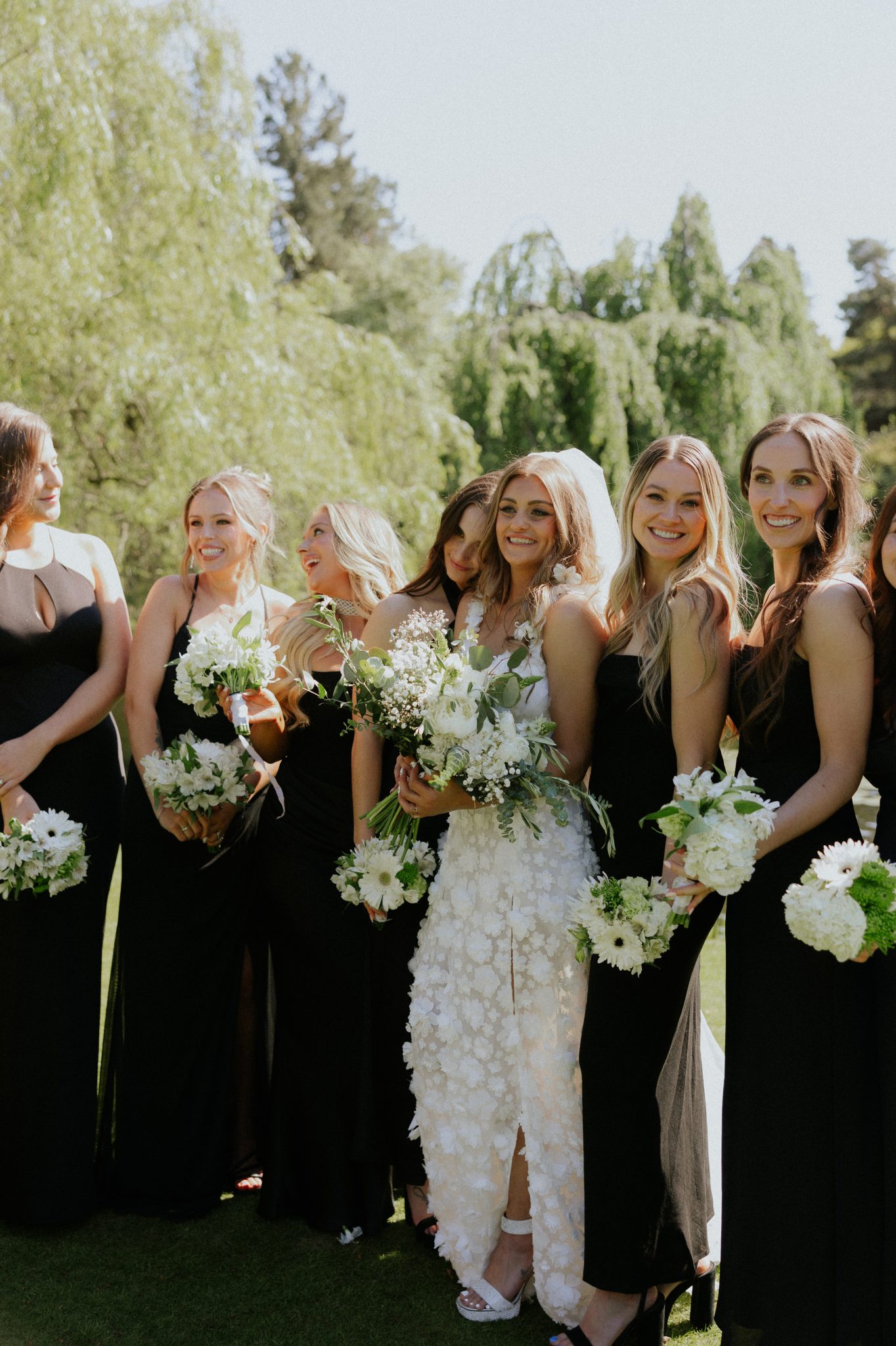 Bride with her bridesmaids dressed in black, holding white bouquets against a lush green backdrop at a Black and White Vancouver Wedding.