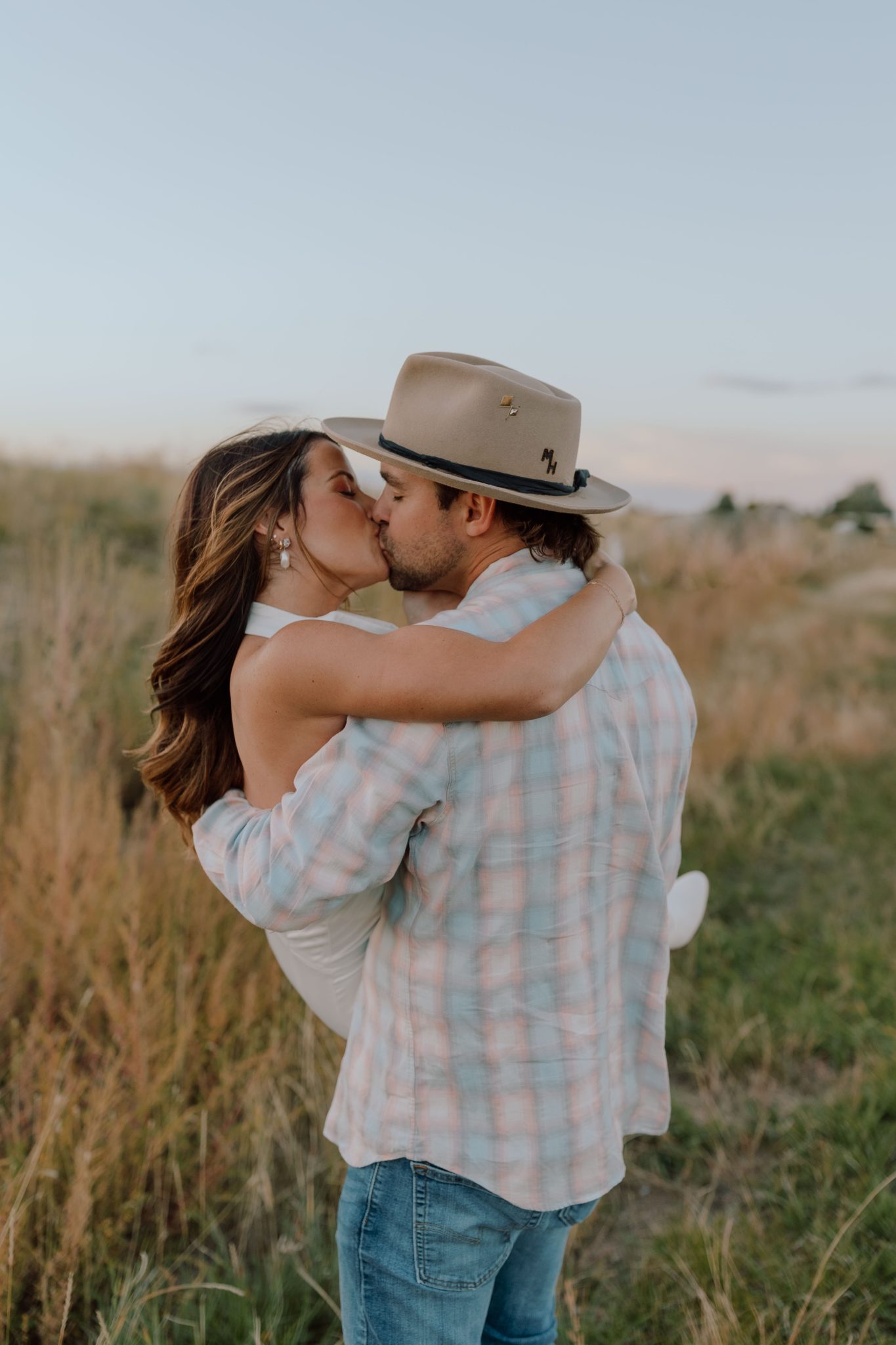 A romantic moment as the groom lifts his fiancée in a field, both wearing hats and cowboy boots, embodying the rustic charm of their Western Fall Engagement Session in Colorado.