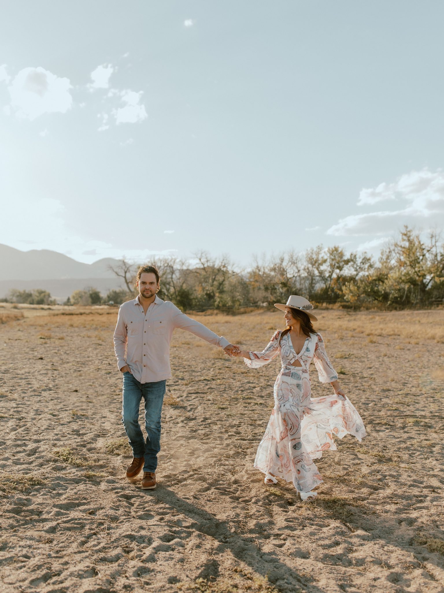 A couple holding hands and playfully walking in an open field with mountains in the background, highlighting their carefree love during their Western Fall Engagement Session in Colorado.