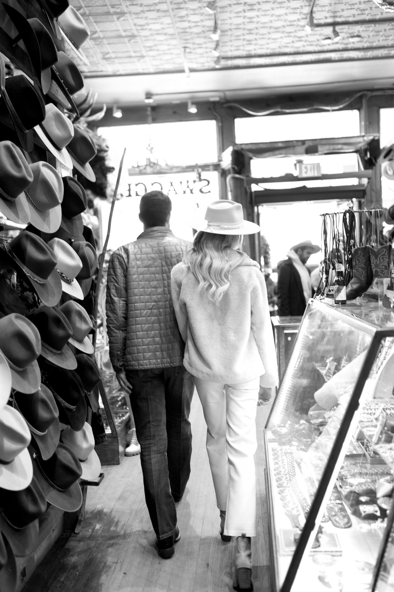 Black and white image of a couple walking through a rustic hat shop in Aspen during their winter engagement session.