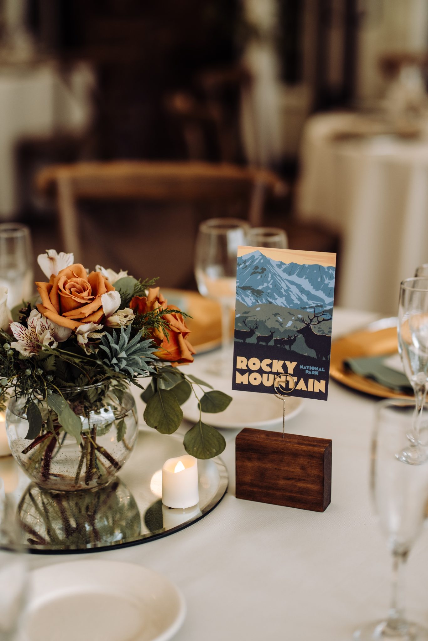 A close-up of a table centerpiece at a Colorado wedding at Tapestry House, featuring vibrant orange and cream flowers, Rocky Mountain-themed table cards, and soft candlelight.