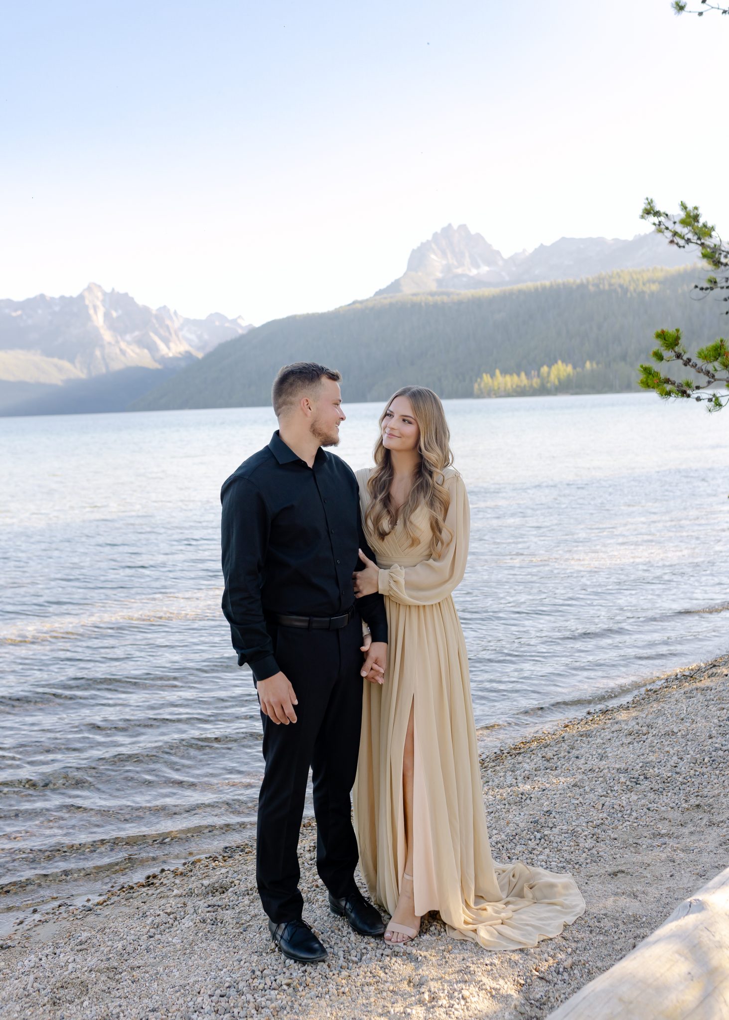A couple standing by a serene lakeside, with mountains in the background during their Promise Me Film and Photo summer engagement session.