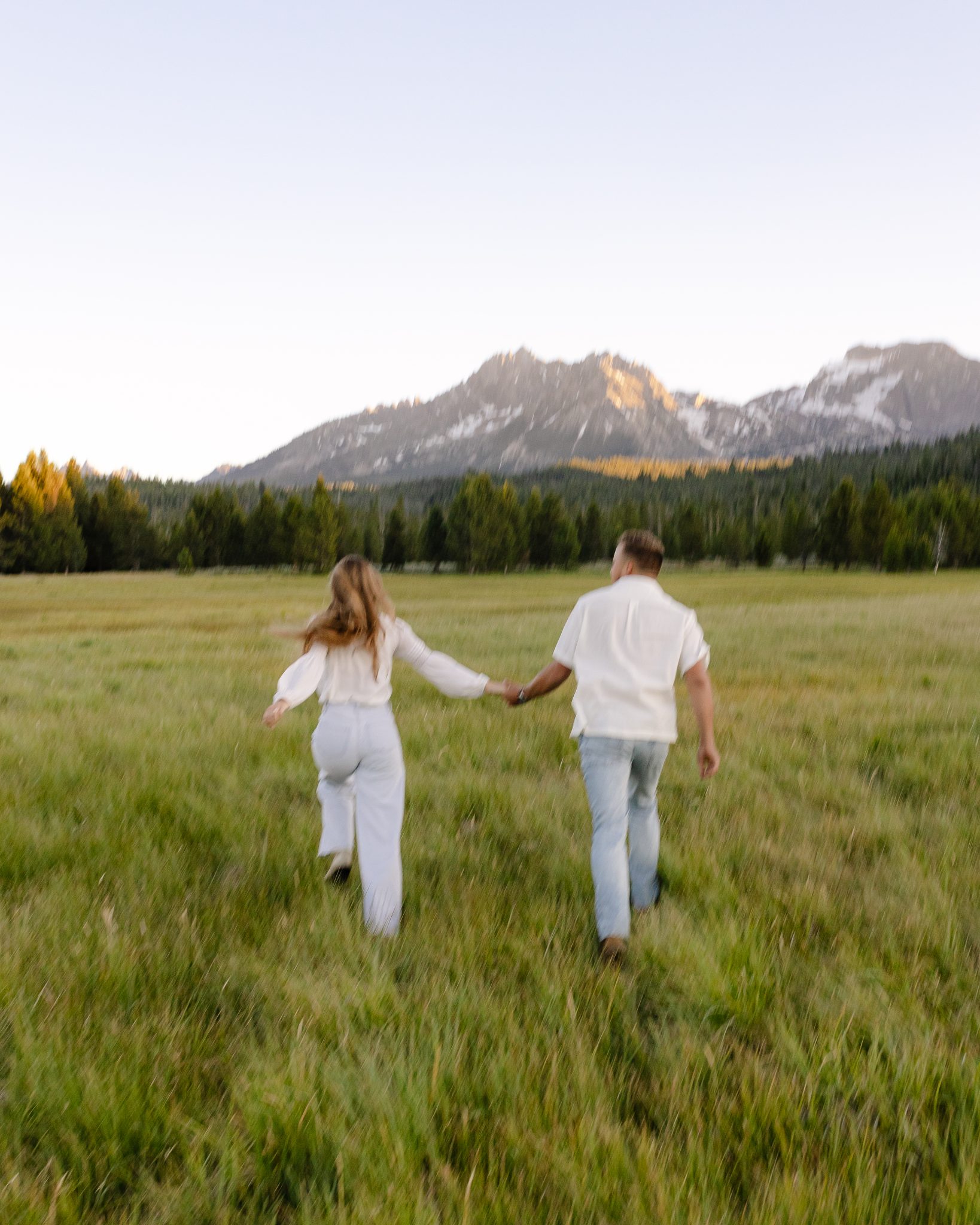 Playful couple walking through a green field, framed by towering mountains in the background, during their Promise Me Film and Photo summer engagement shoot.
