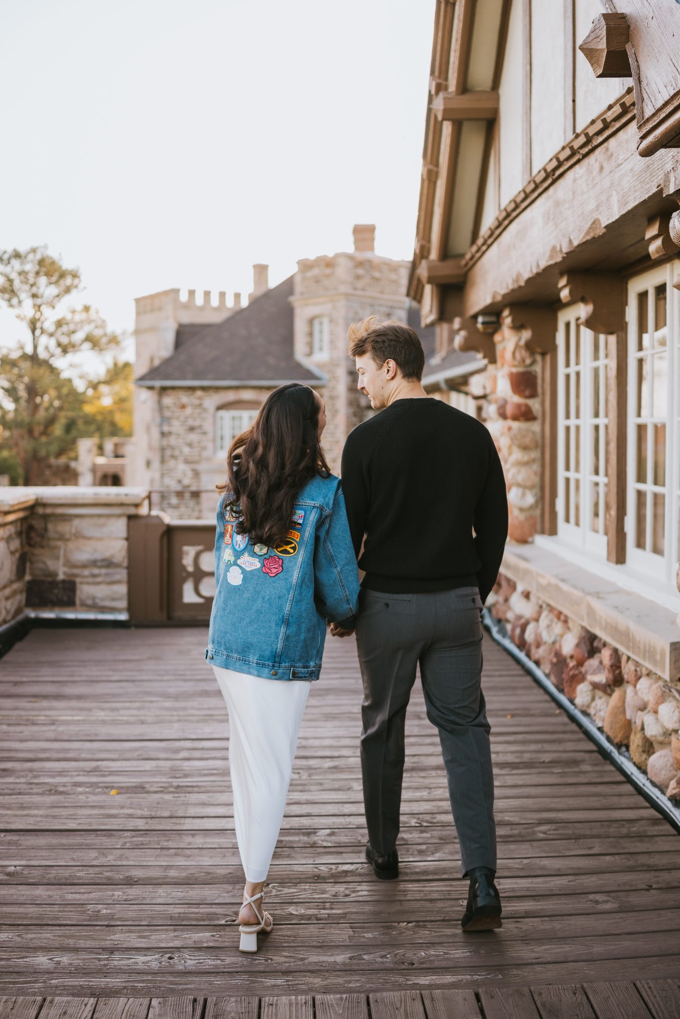 The couple walks hand-in-hand on the elegant wooden balcony of Highlands Ranch Mansion during their fall engagement session.