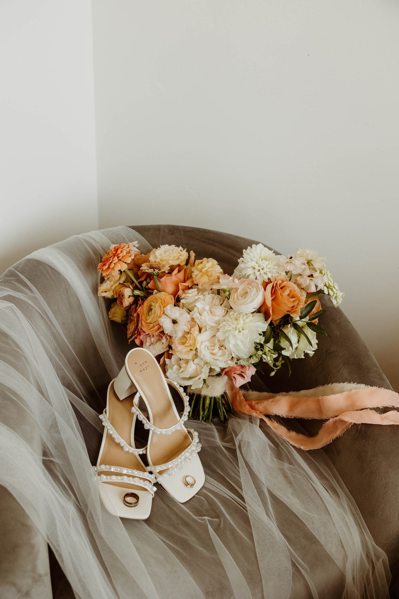 Close-up of the bride’s elegant shoes adorned with pearls, paired with a soft orange and cream floral bouquet resting on a chair, highlighting the soft orange wedding color palette.