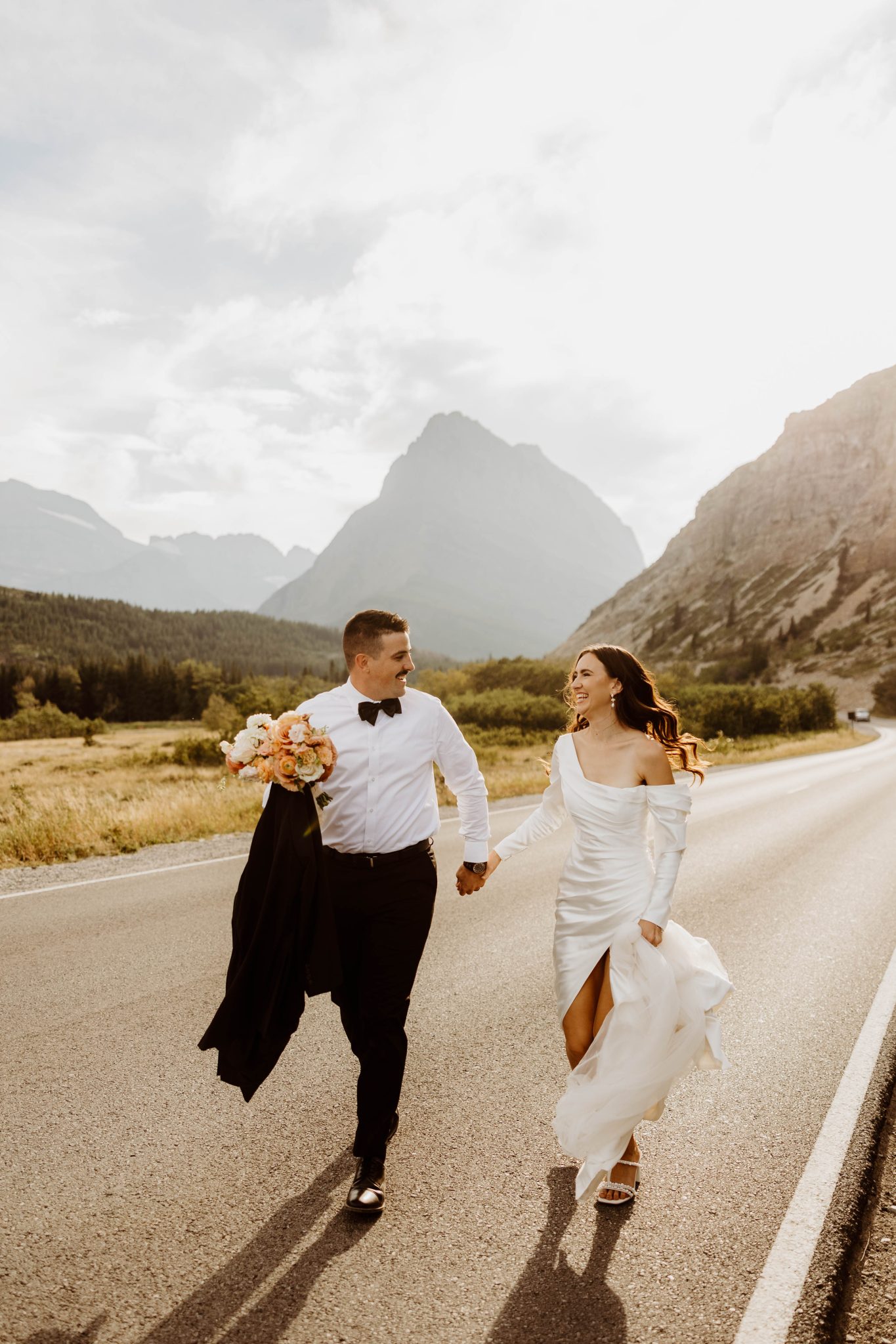 The couple smiles as they walk down a scenic road, the bride holding a bouquet with soft orange hues, capturing the warm tones of the soft orange wedding color palette.