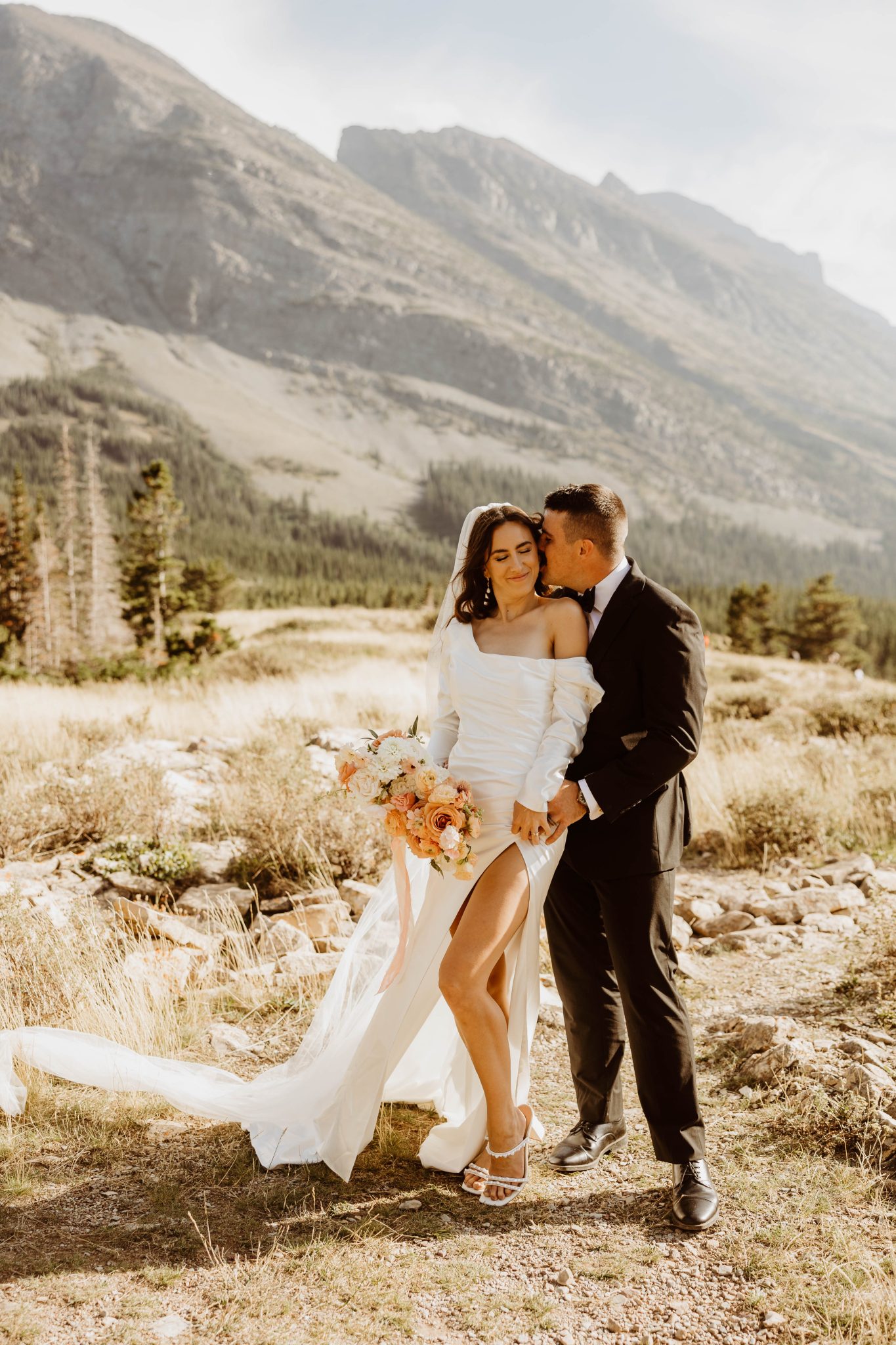 Romantic moment of the bride and groom with a stunning mountain backdrop, showcasing the bride’s soft orange floral bouquet and elegant off-shoulder gown, fitting the soft orange wedding color palette.
