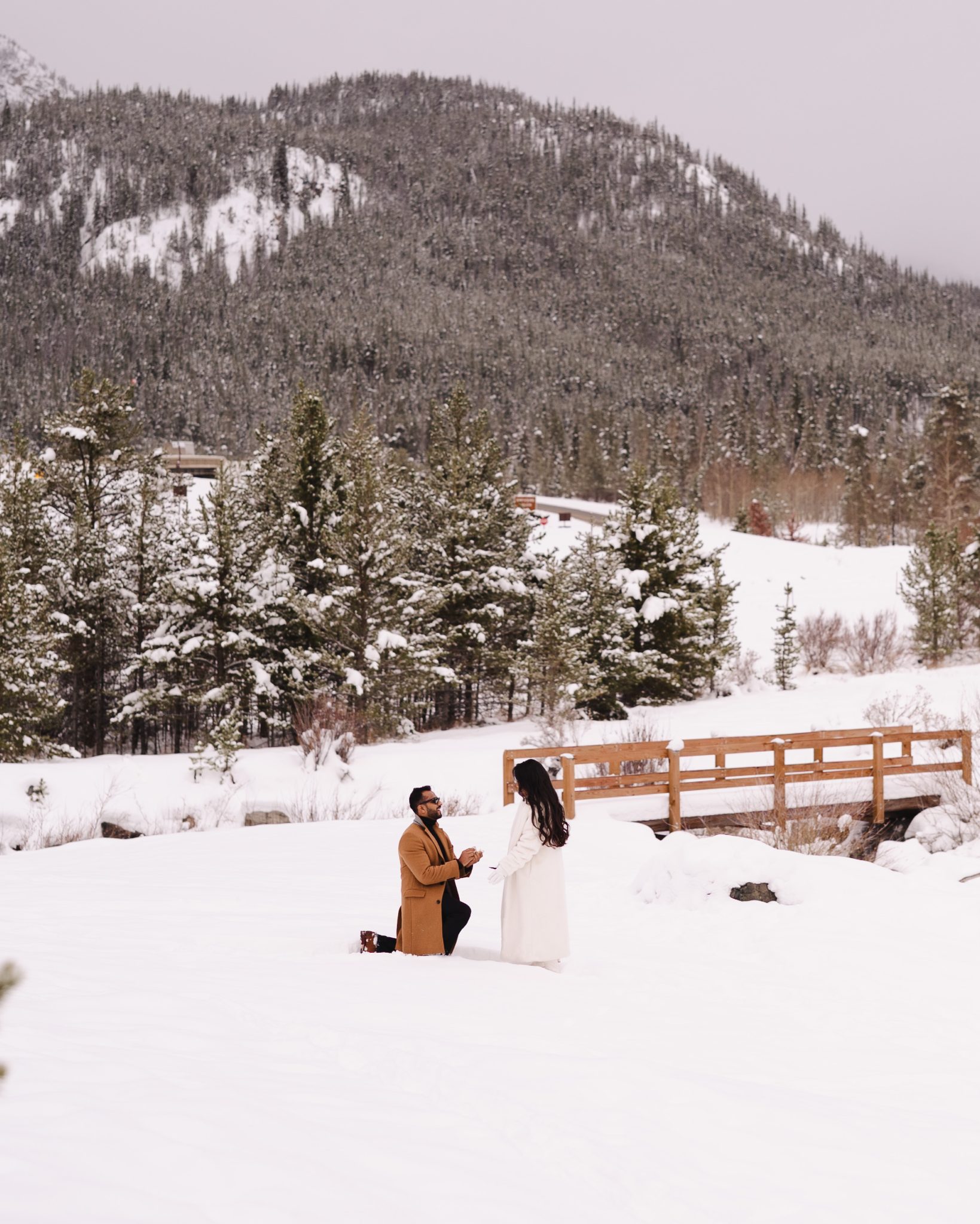 A wide shot of a groom-to-be kneeling in the snow proposing, set against a backdrop of snowy trees and a wooden bridge during a Colorado surprise proposal.