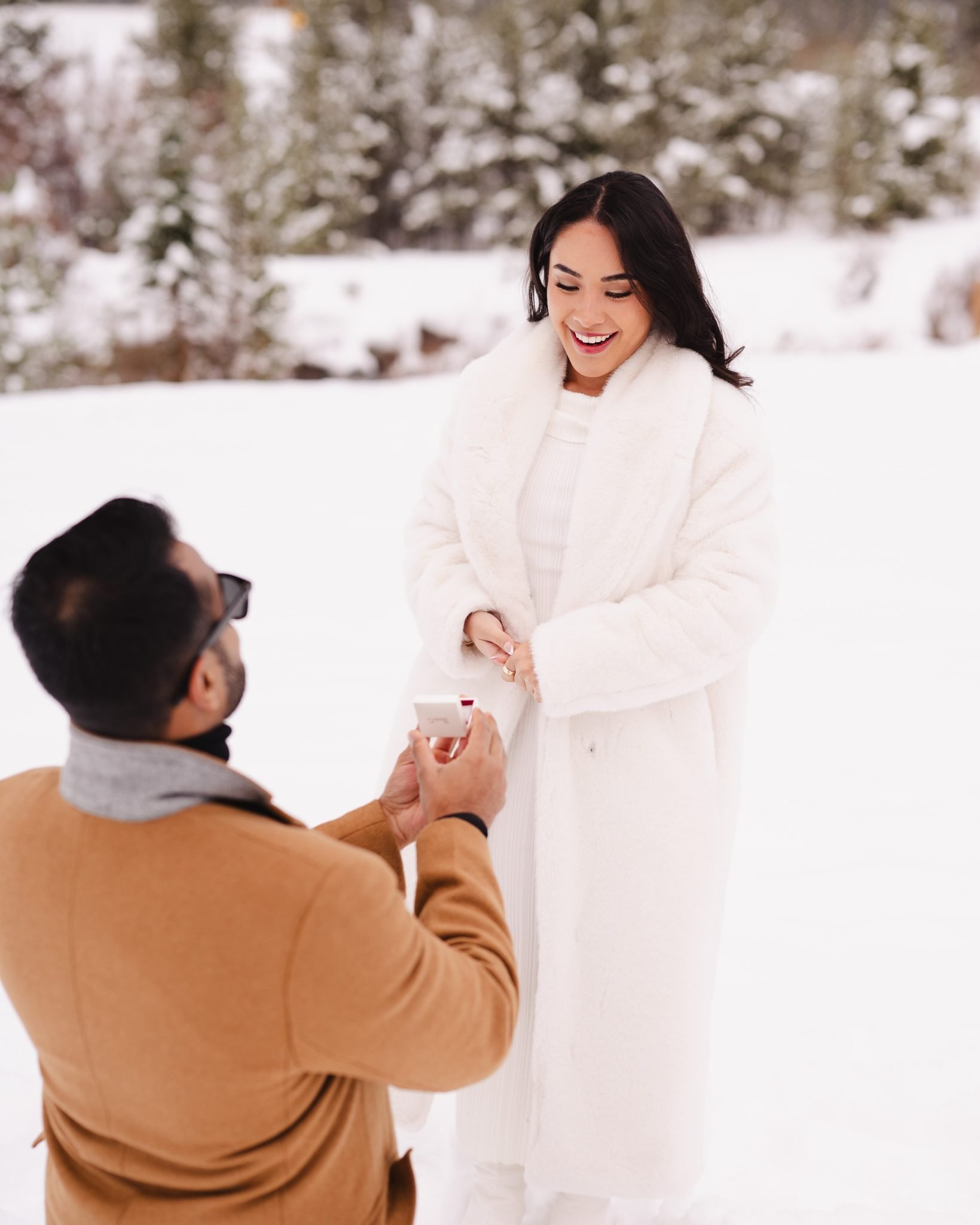 A groom-to-be kneels in the snow with a ring box during a magical Colorado surprise proposal, surrounded by a winter wonderland.