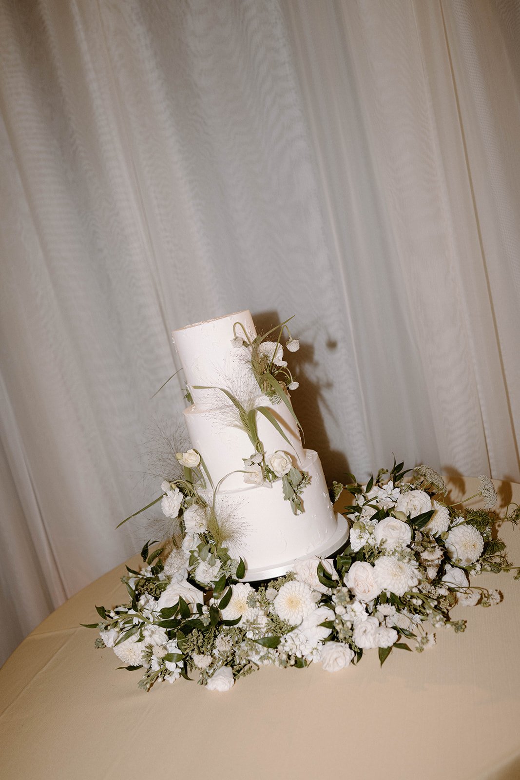 A minimalist three-tiered white wedding cake adorned with delicate greenery and florals at an elevated and neutral summer Aspen wedding.