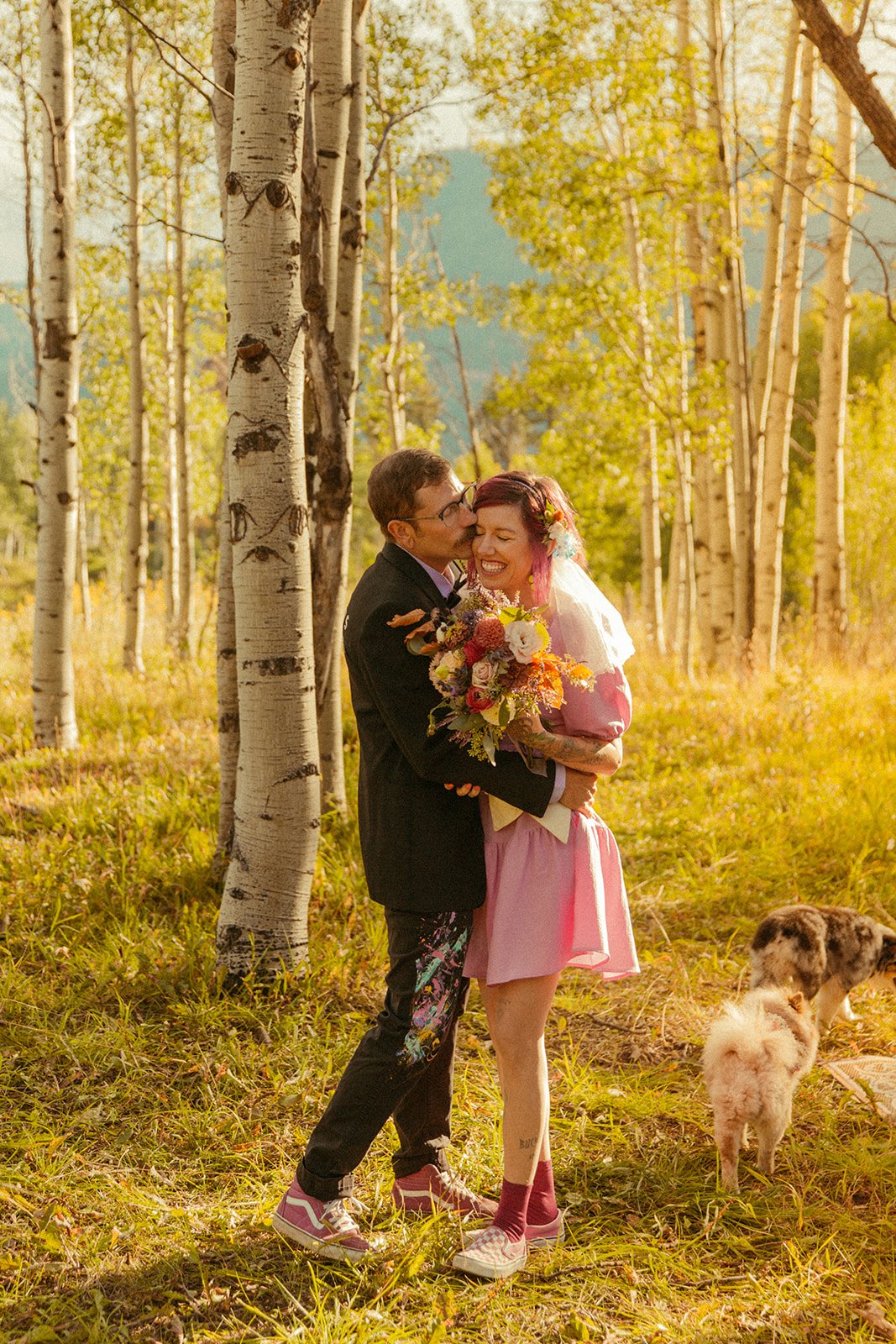 A joyful couple sharing an embrace in a forest of aspen trees during their Hanna Irene Motion Pictures Durango Elopement, with pops of colorful flowers and playful attire.