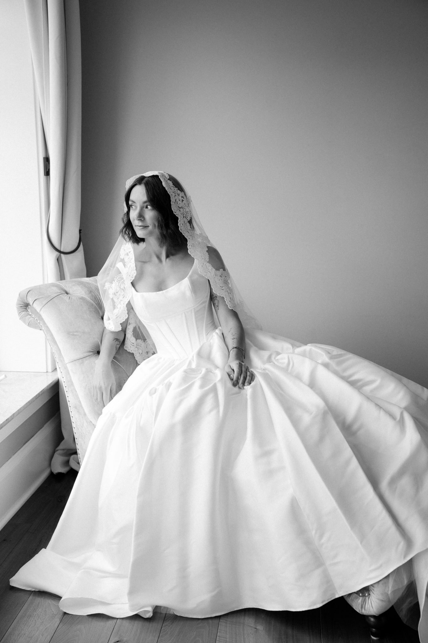 The bride sits gracefully in a vintage chair wearing a timeless lace veil and satin gown during a Summer British Columbia wedding.