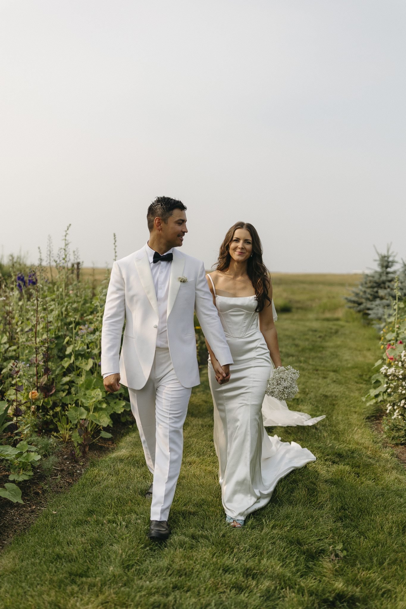 The bride and groom walk hand-in-hand through a lush garden, showcasing elegance and charm at The Gathered weddings.