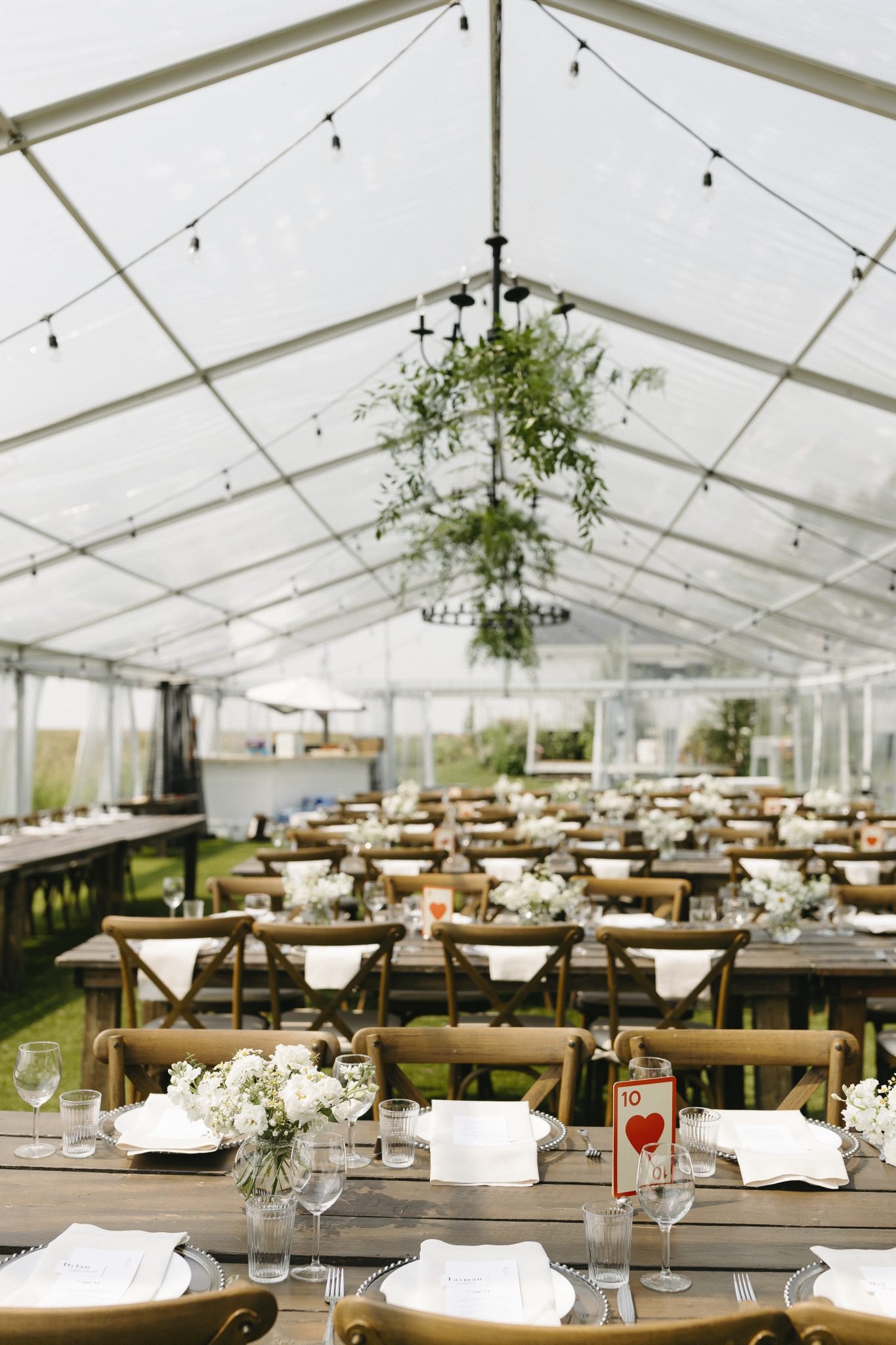A beautifully styled tent reception features wooden tables, white florals, and greenery chandeliers at The Gathered weddings.