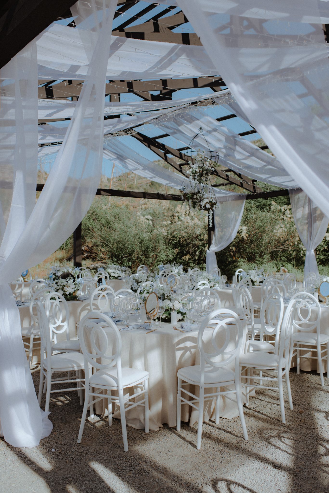 An outdoor reception setup featuring white draped ceilings, white chairs, and floral centerpieces for a sophisticated white and blue wedding.