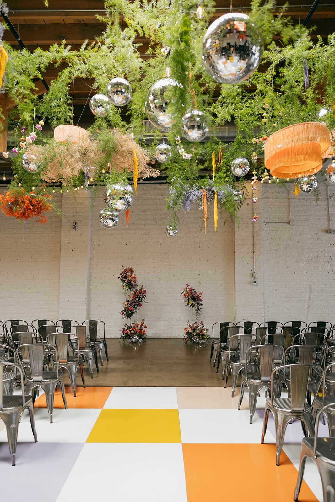 A ceremony setup featuring colorful floral installations, metal chairs, and bold geometric flooring, showcasing a unique and stylish modern wedding design.