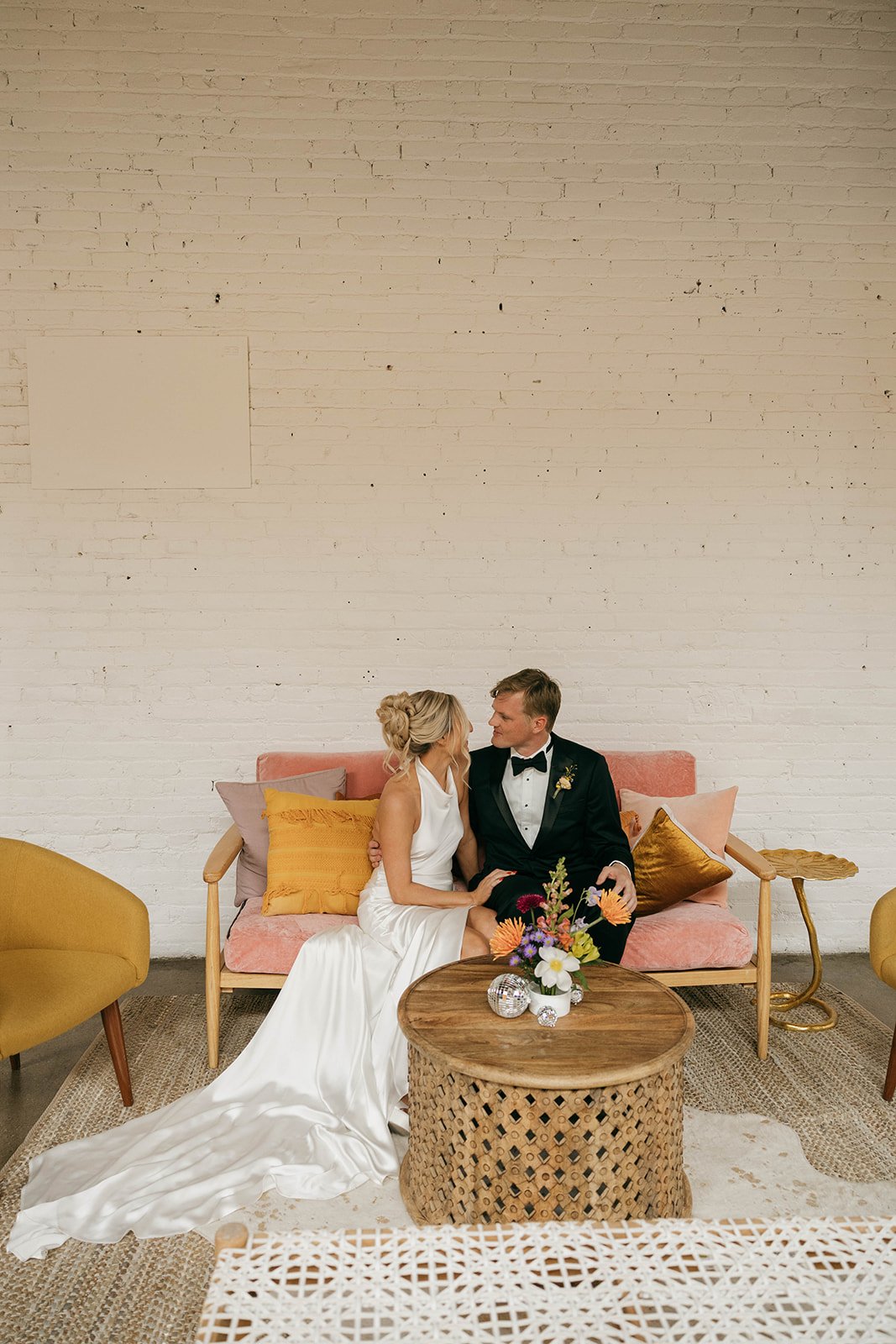 The bride and groom share an intimate moment on a pink and mustard lounge setup, complete with cozy pillows and small floral accents, elevating the style of a modern wedding.