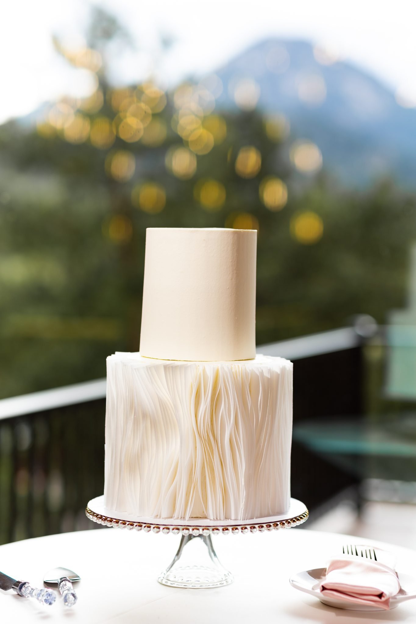 A modern two-tier white wedding cake with textured details, displayed with mountain views in the background at an Estes Park wedding.