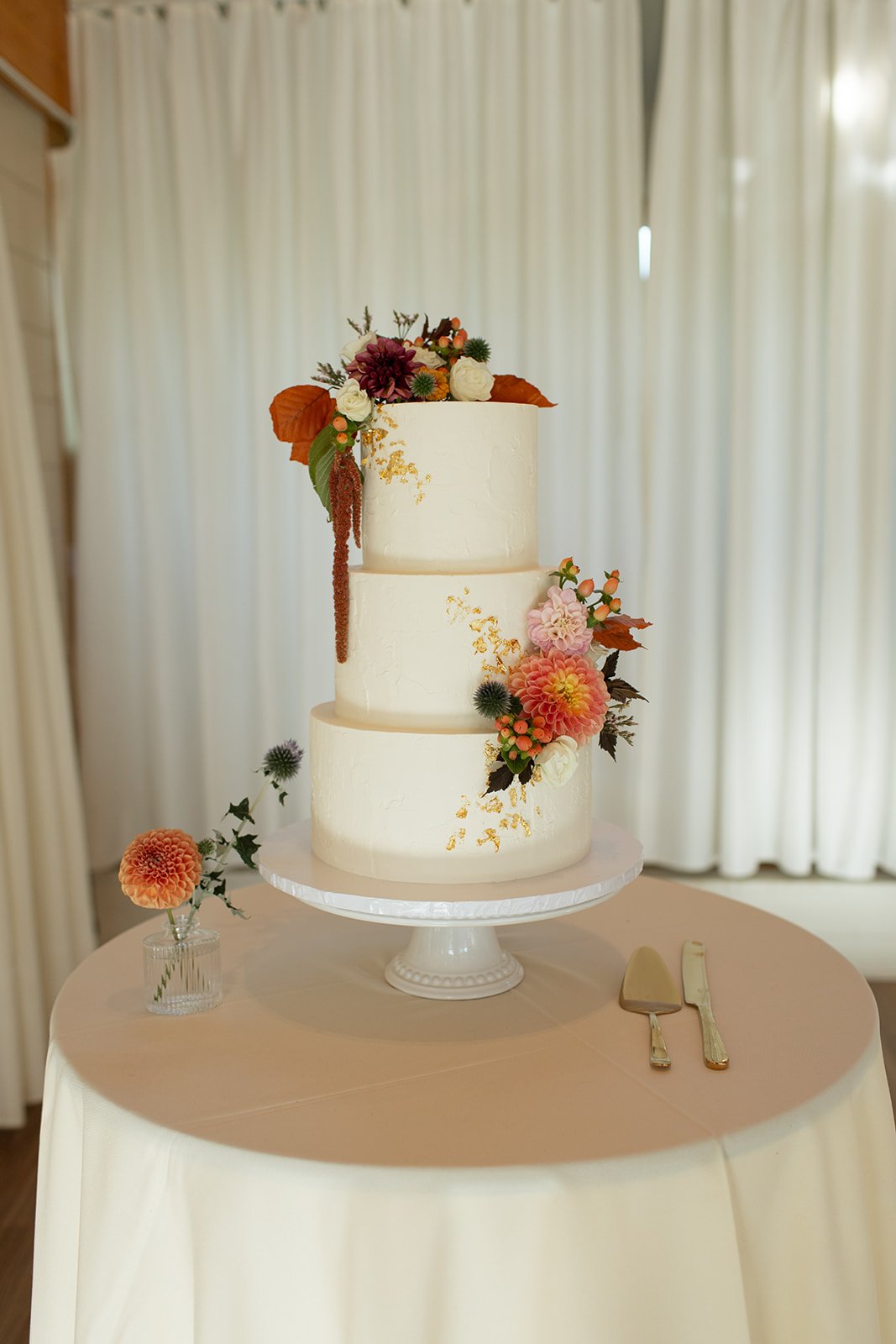 Three-tier wedding cake adorned with fall-inspired floral decorations, featuring gold accents at a Fall Alberta wedding.